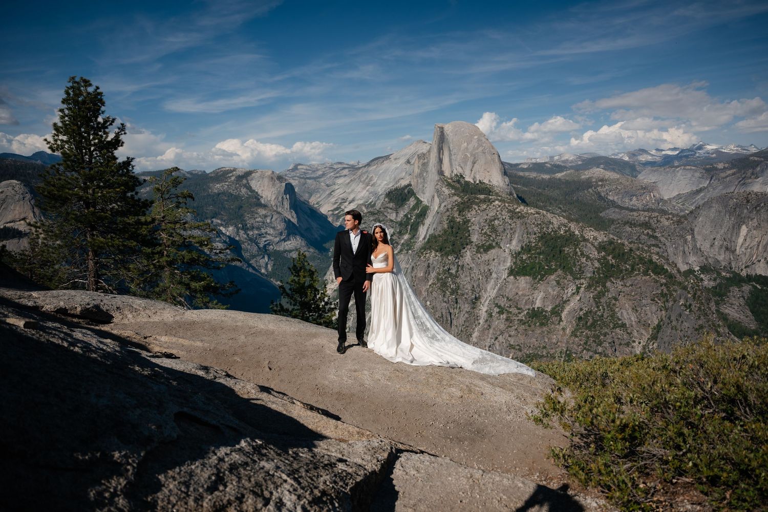 Bride and groom stand on rocky overlook at Glacier Point, Yosemite, with Half Dome and mountain range behind them.