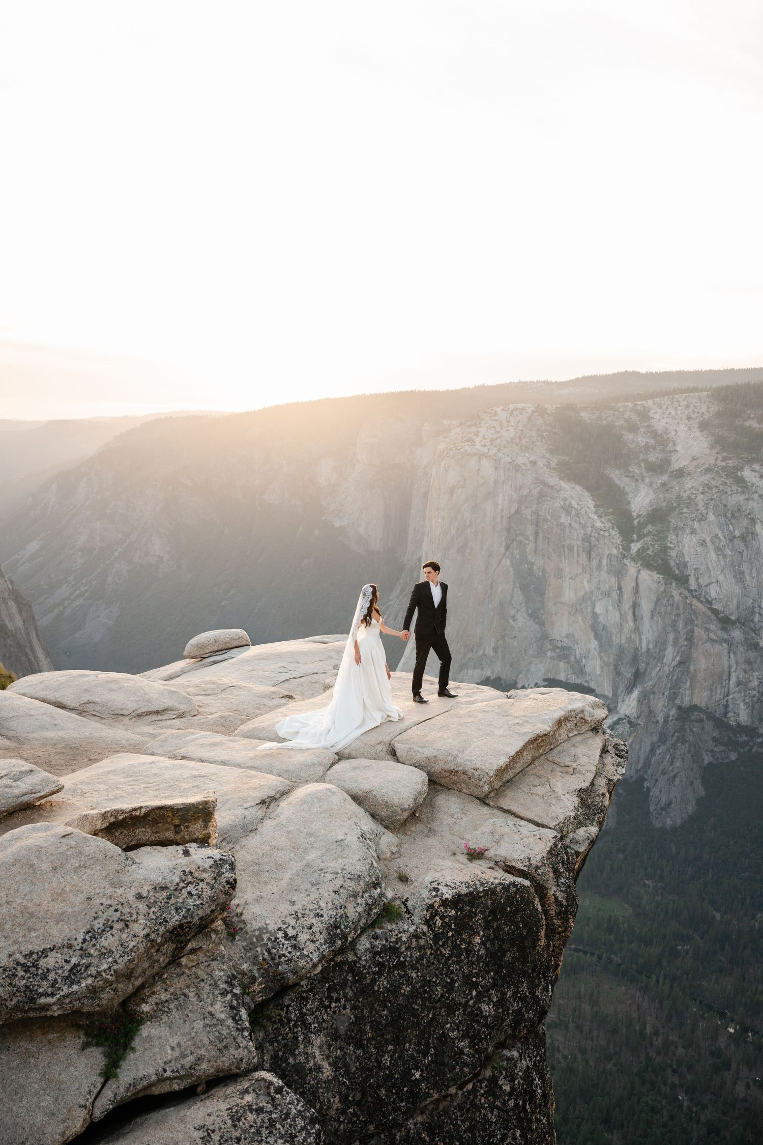 Bride and groom hold hands on dramatic rocky mountain cliff edge at sunset in Yosemite National Park.