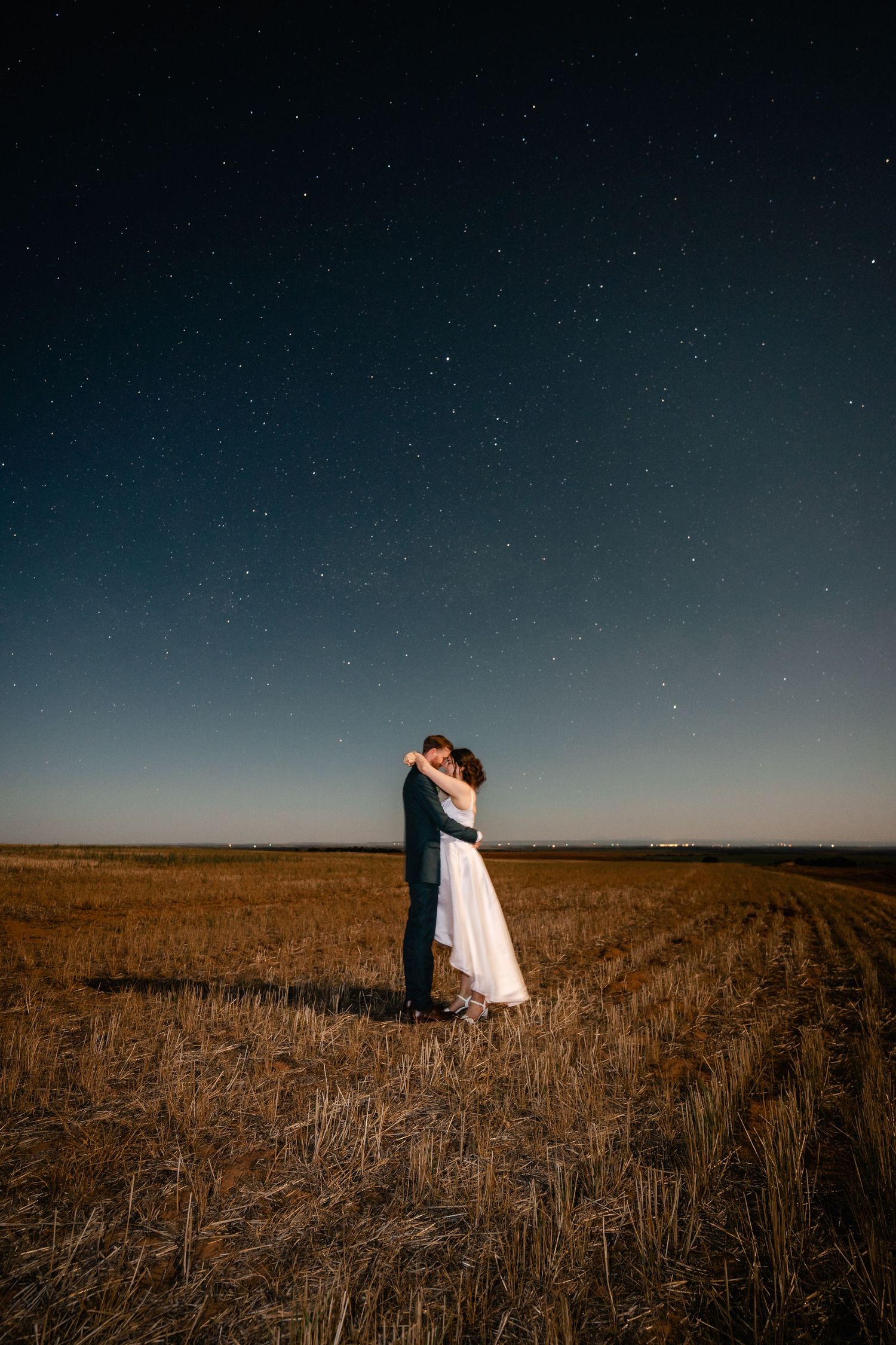 A romantic wedding photo at night showing silhouettes against a starry sky in an open field.