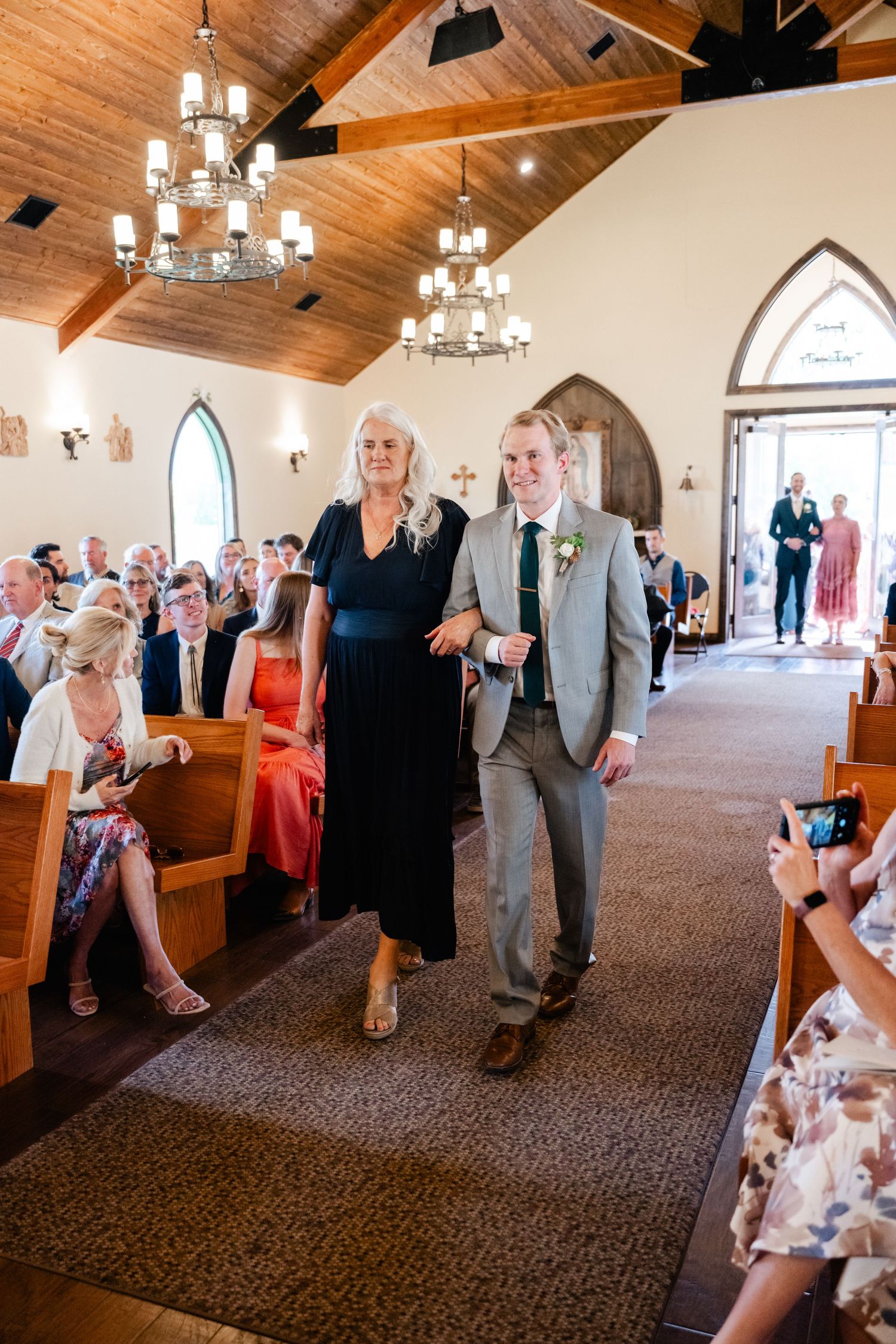 Wedding guests mingle in a church hall with wooden ceiling beams and chandeliers.