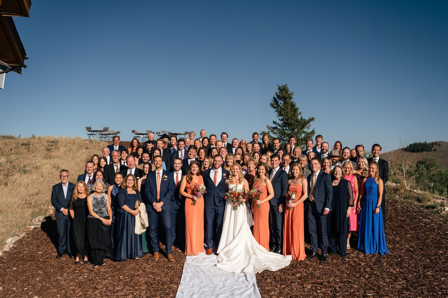 Large wedding party gathered outdoors with mountain landscape and blue sky backdrop.