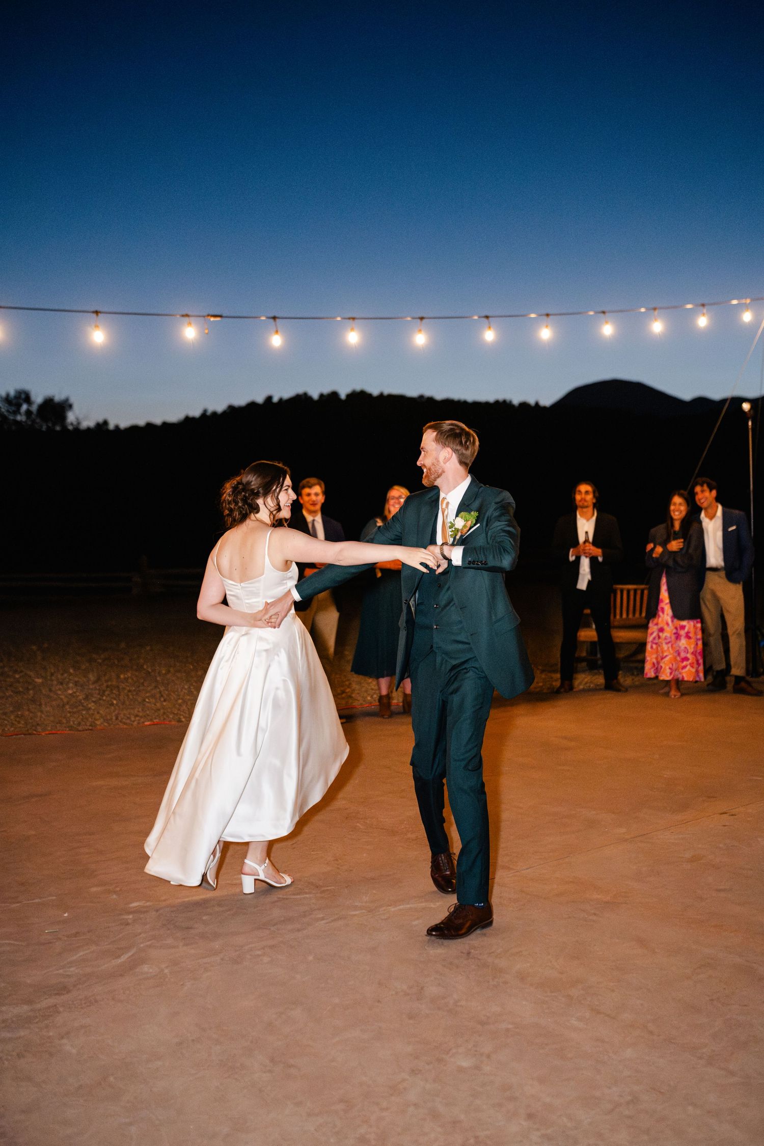 Newlyweds dancing under string lights at dusk during their outdoor reception.