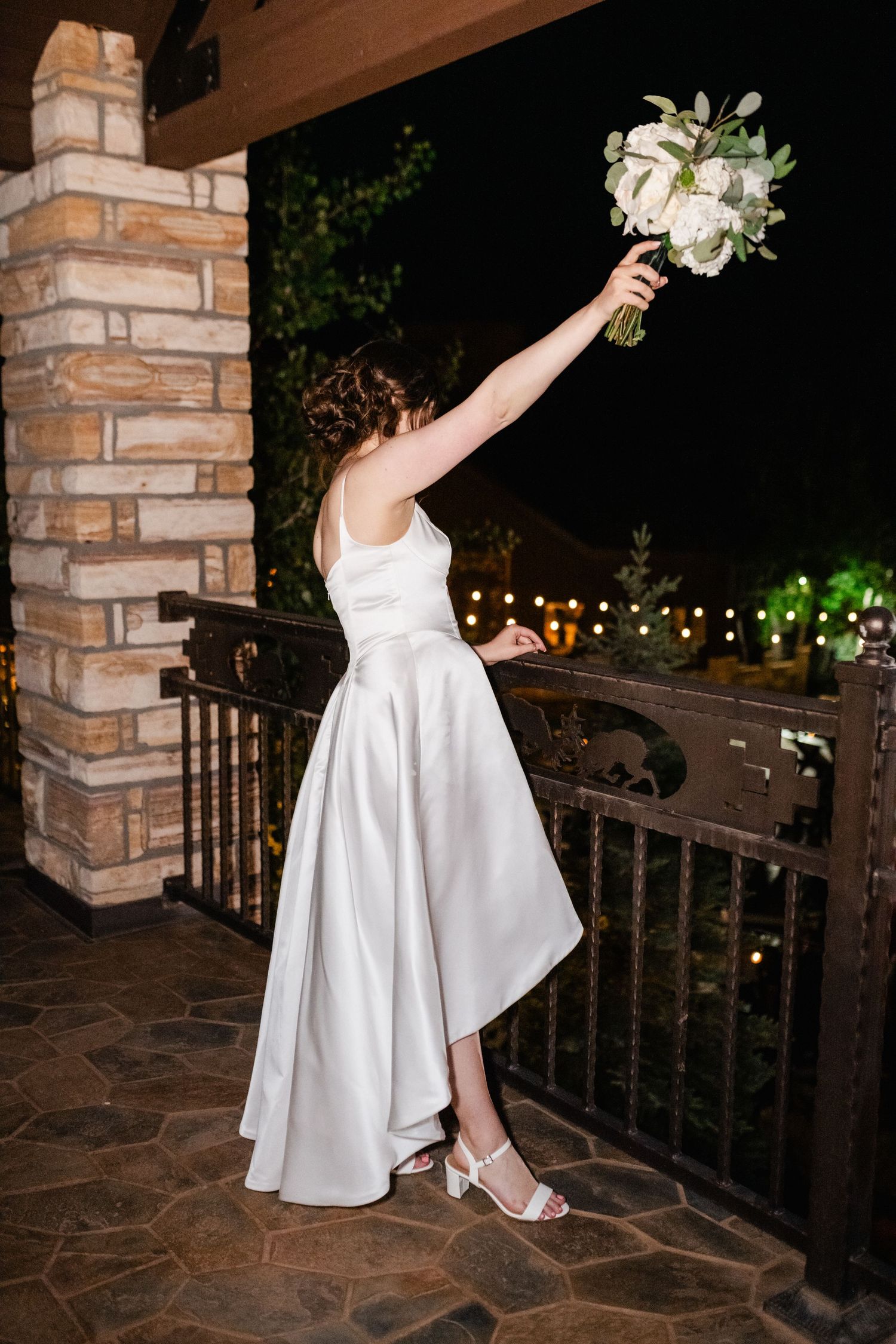 A bride poses with her bouquet on a stone terrace with twinkling lights in the background.