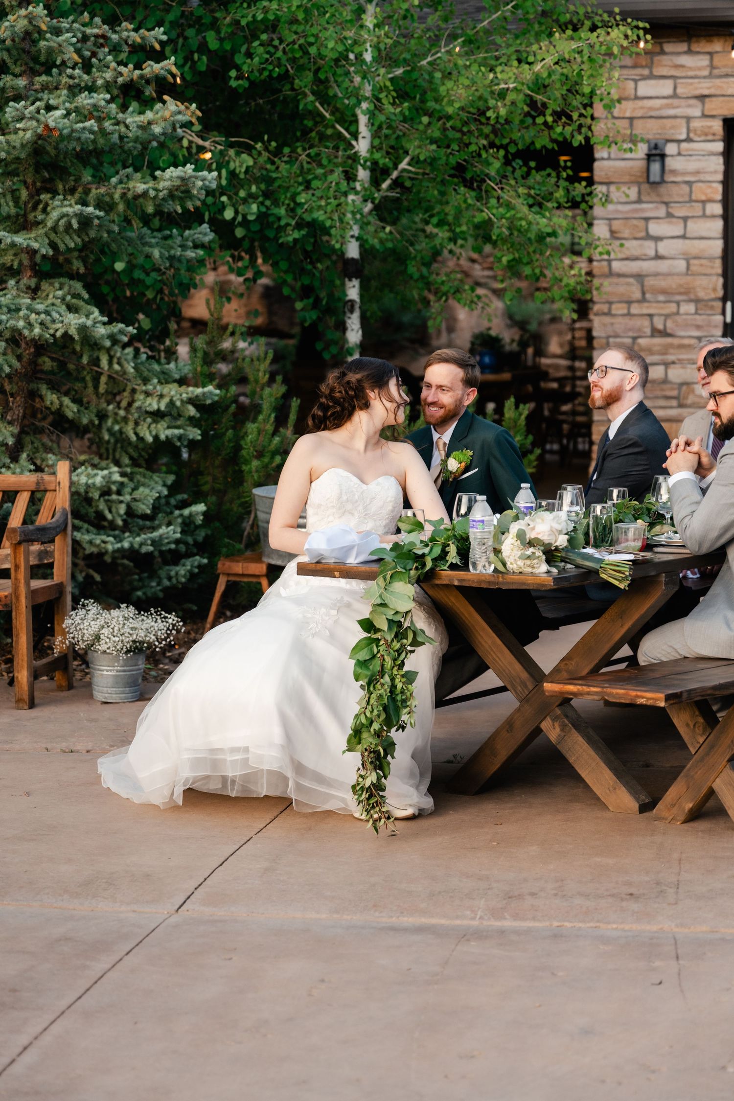 Wedding reception dining setup with rustic wooden picnic tables and greenery decorations on a stone patio.