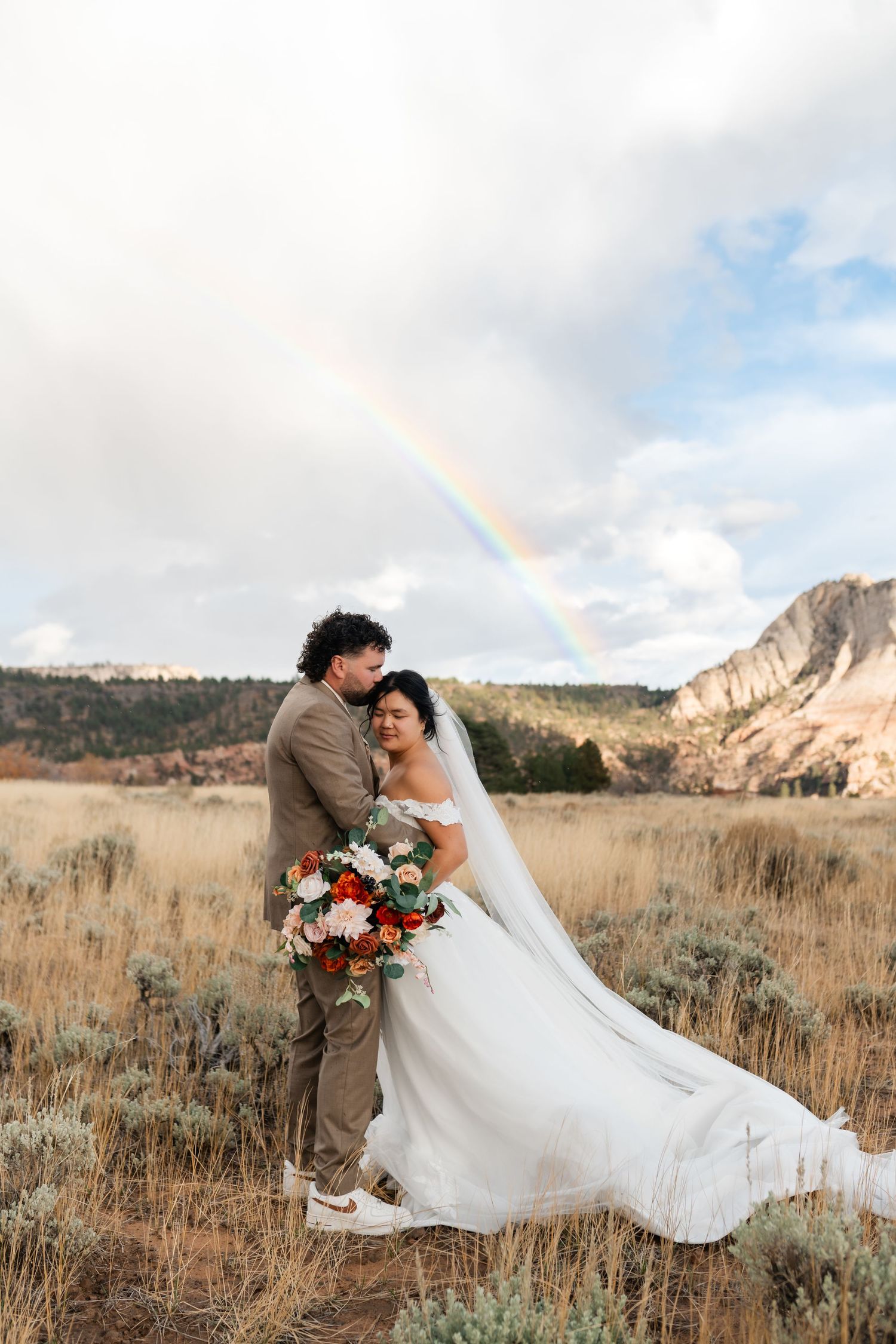 Wedding couple poses in field with dramatic mountain landscape and rainbow in cloudy sky.
