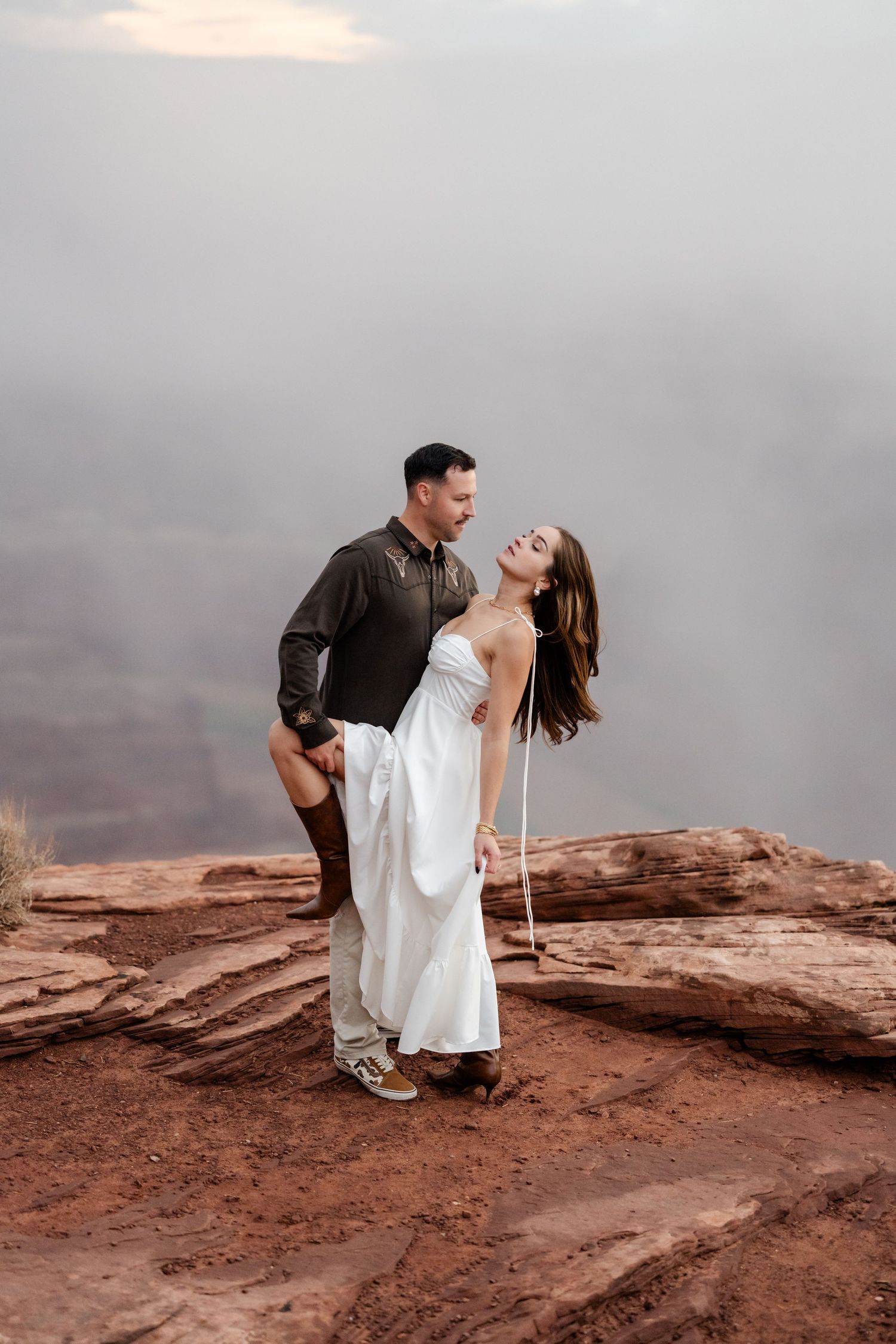 A couple poses dramatically on red rock cliffs during an overcast day.