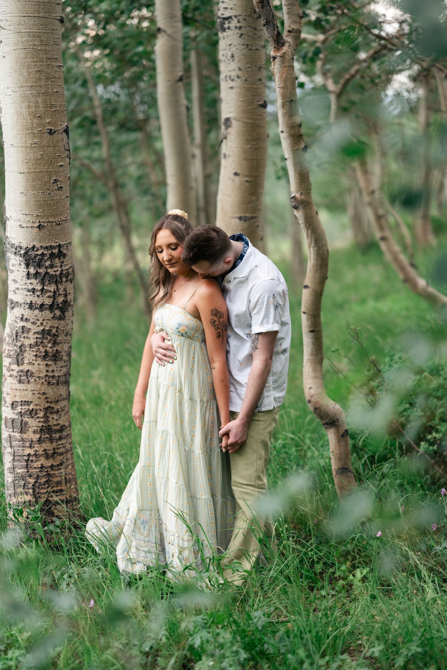 A couple shares an intimate moment among white birch trees in a lush forest setting.