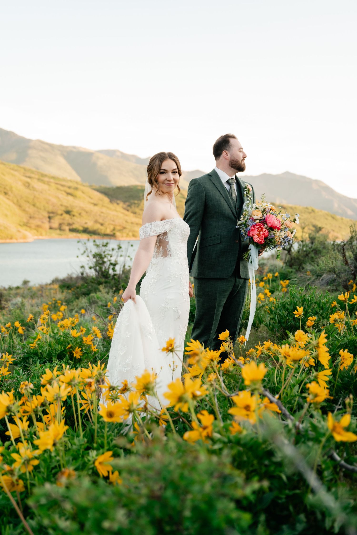 Newlywed couple stands together in a field of vibrant yellow wildflowers with mountains in the background.