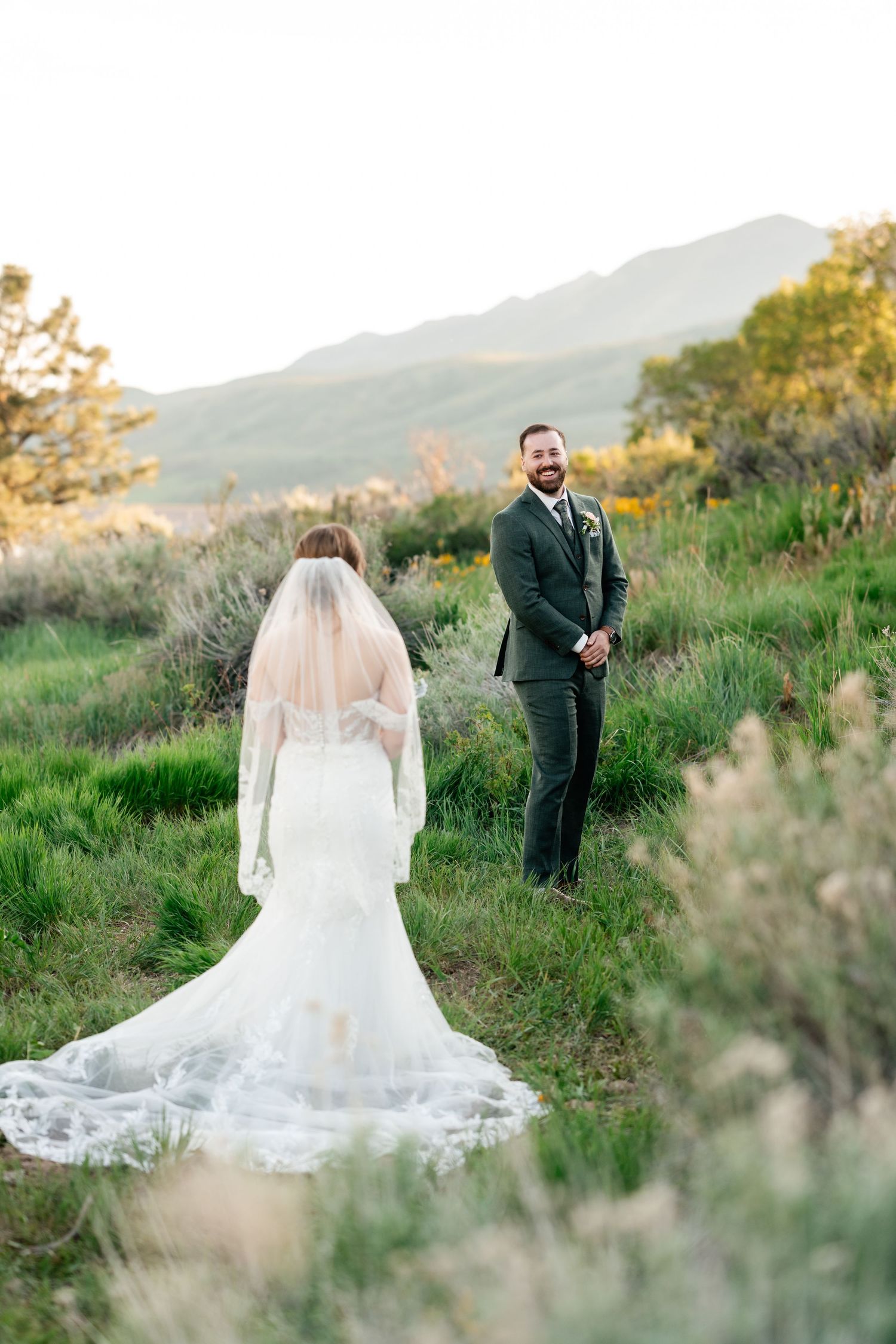 A wedding first look moment captured on a grassy hillside with mountains in the background at sunset.