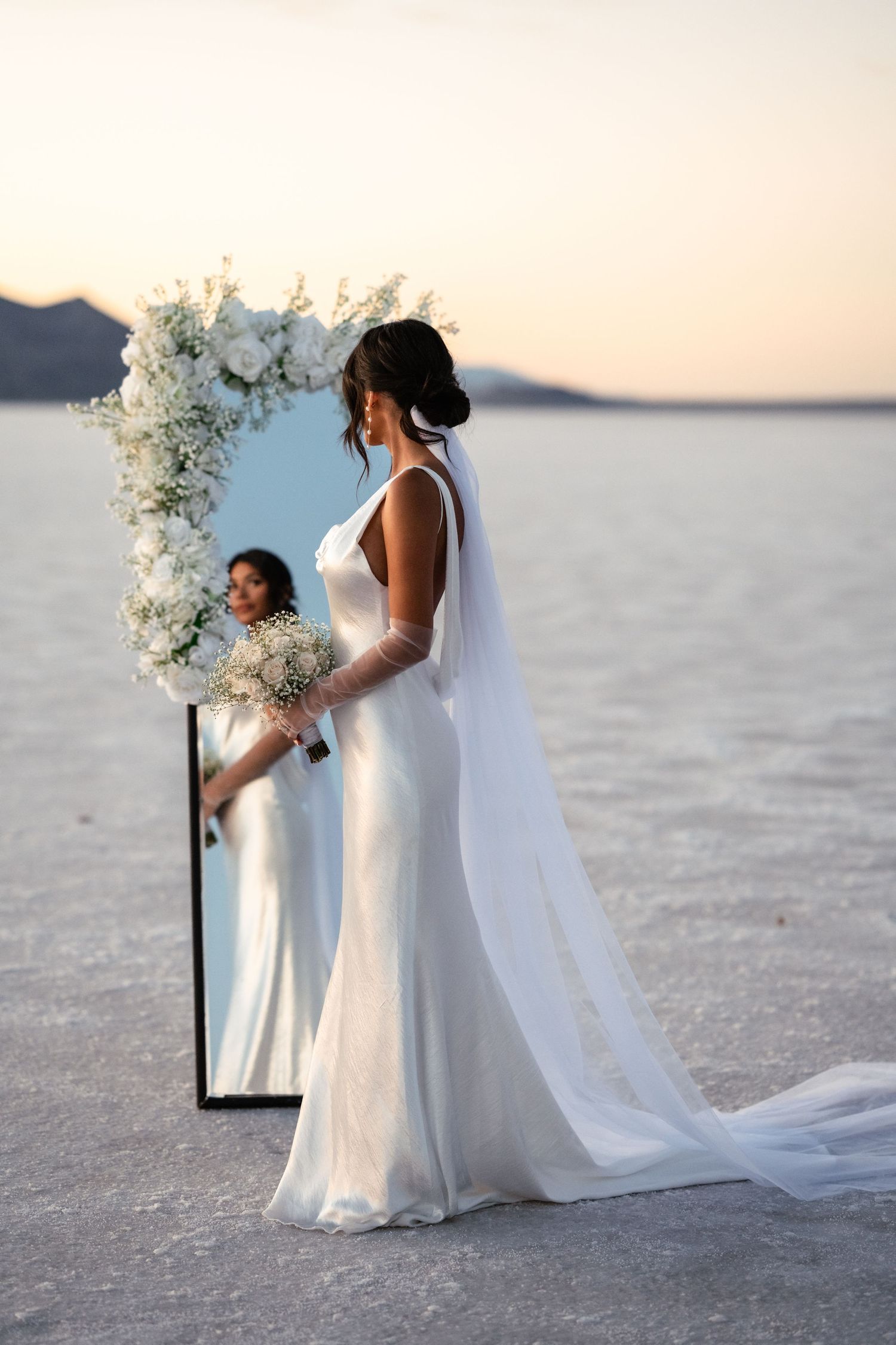 A wedding ceremony setup with white floral arrangements on a salt flat at sunset.