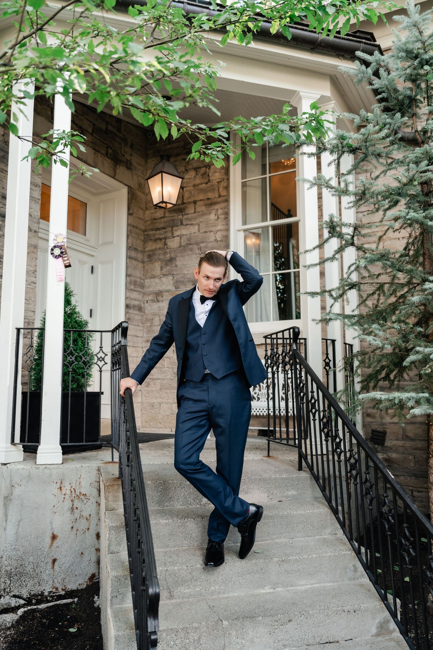 A groom in a navy suit strikes playful poses on stone steps with iron railings.