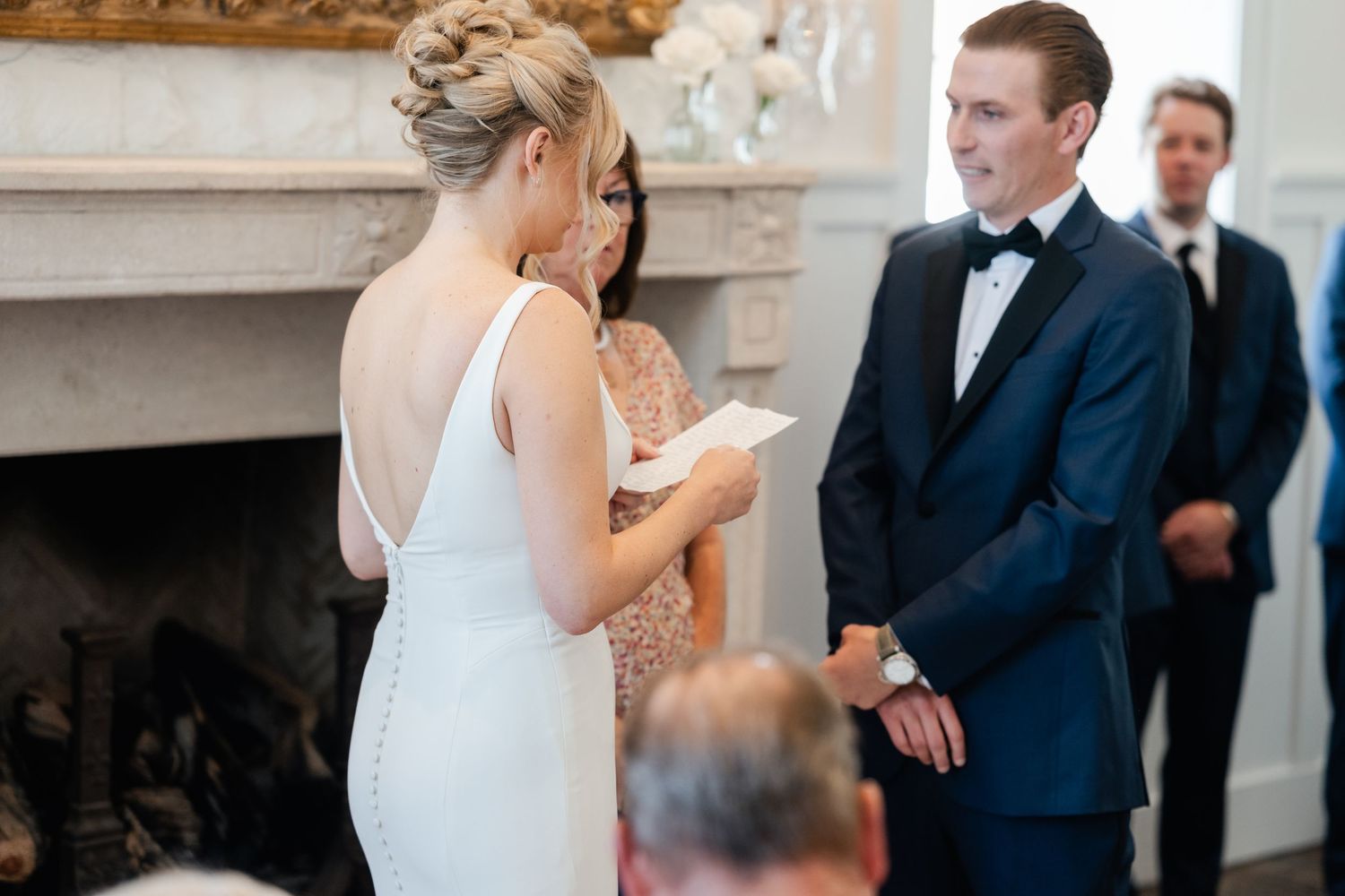A couple exchanges vows during an intimate indoor wedding ceremony beside an ornate fireplace.