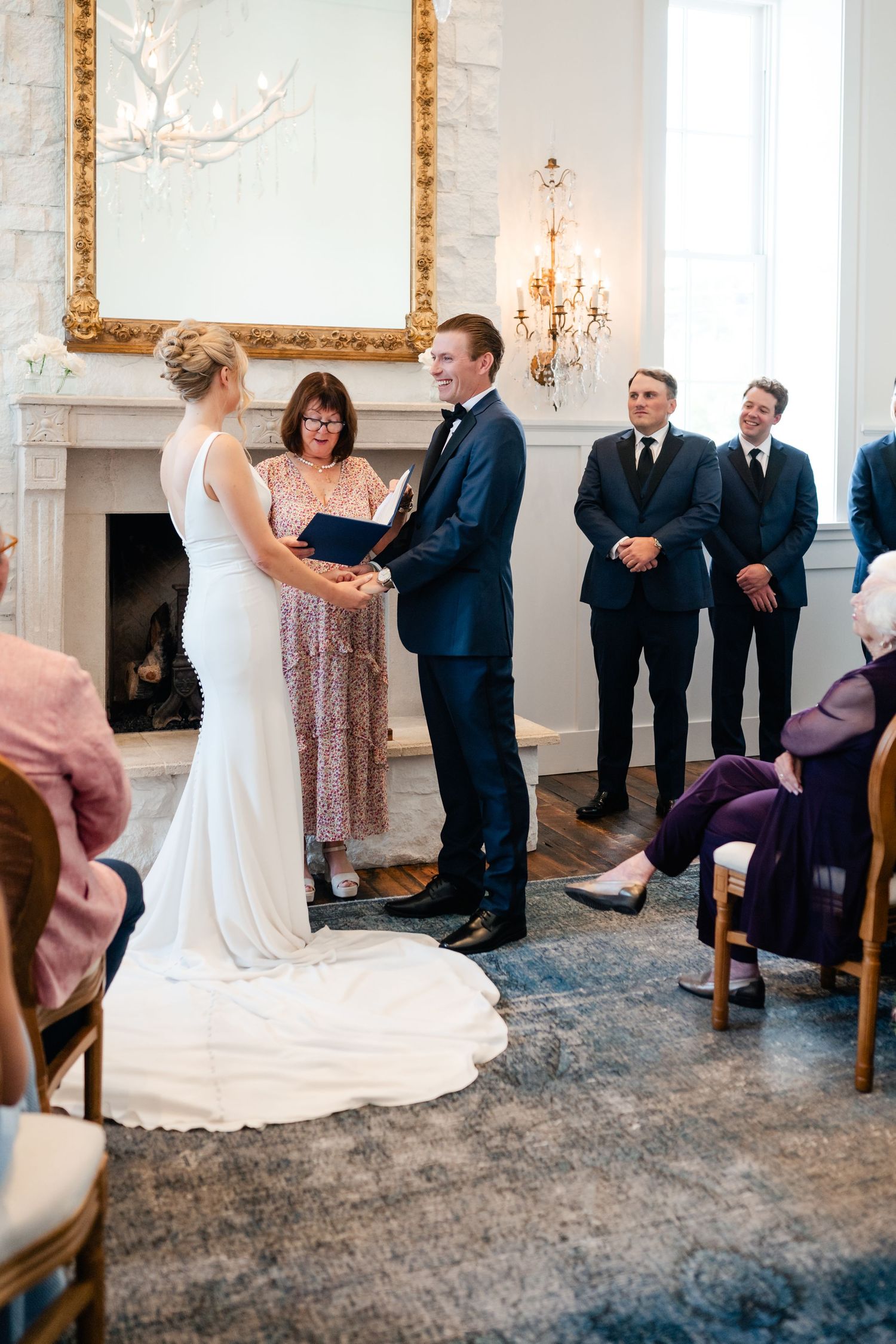 Wedding ceremony participants gathered in an elegant white room with ornate gold mirrors and chandeliers.