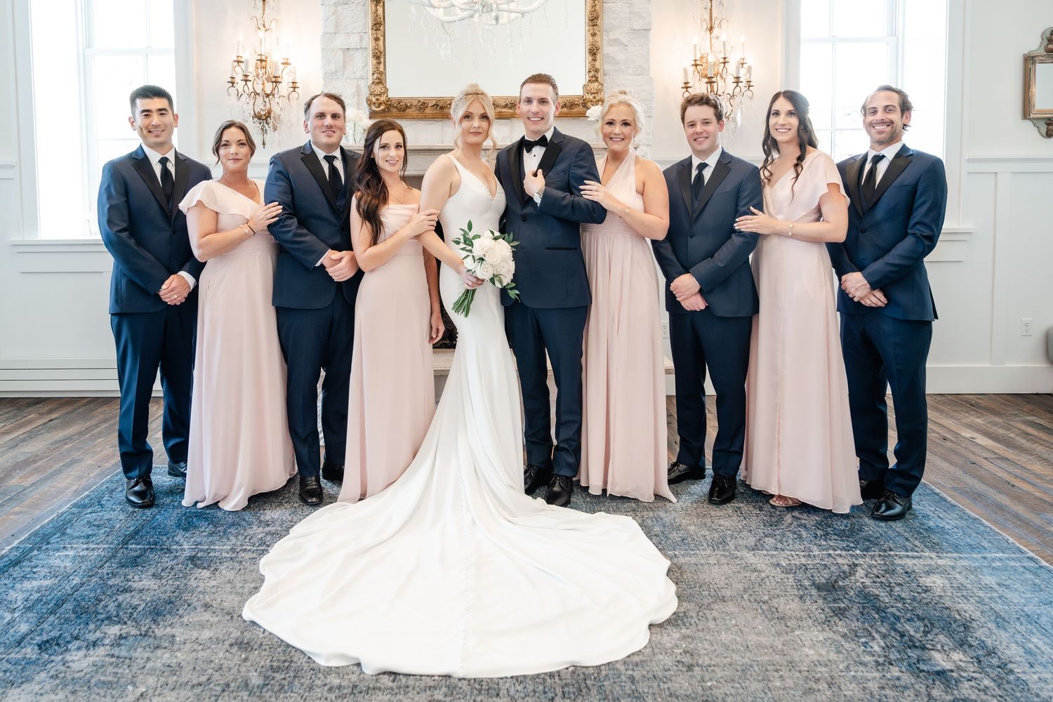 Formal wedding party portrait in bright white chapel with chandeliers and decorative backdrop.