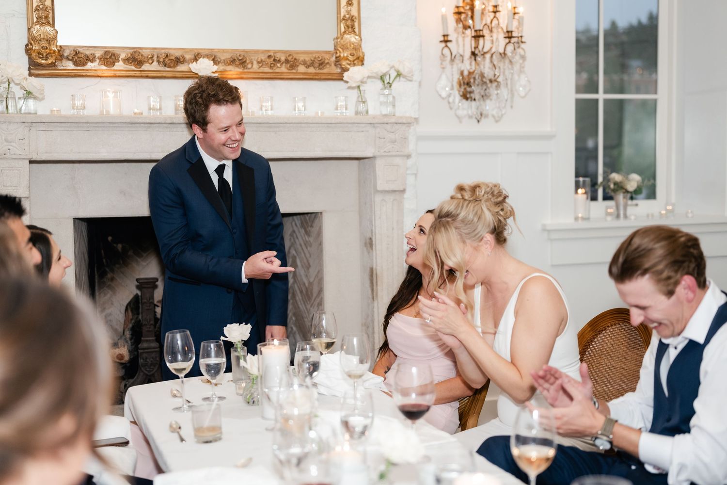Wedding reception guests react during speeches in elegant white room with fireplace and chandelier.