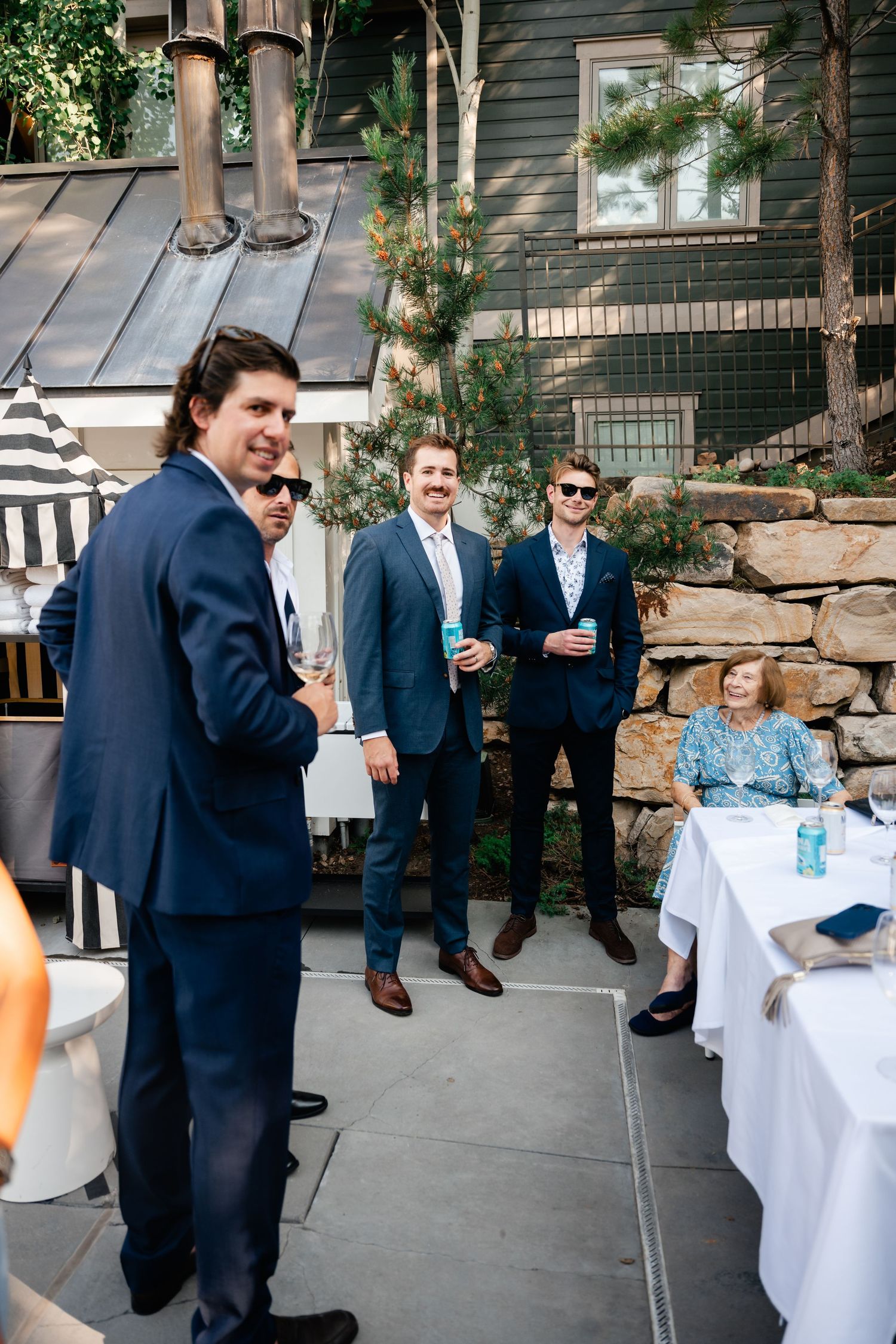 Wedding party members laugh and socialize on an outdoor patio near stone walls.