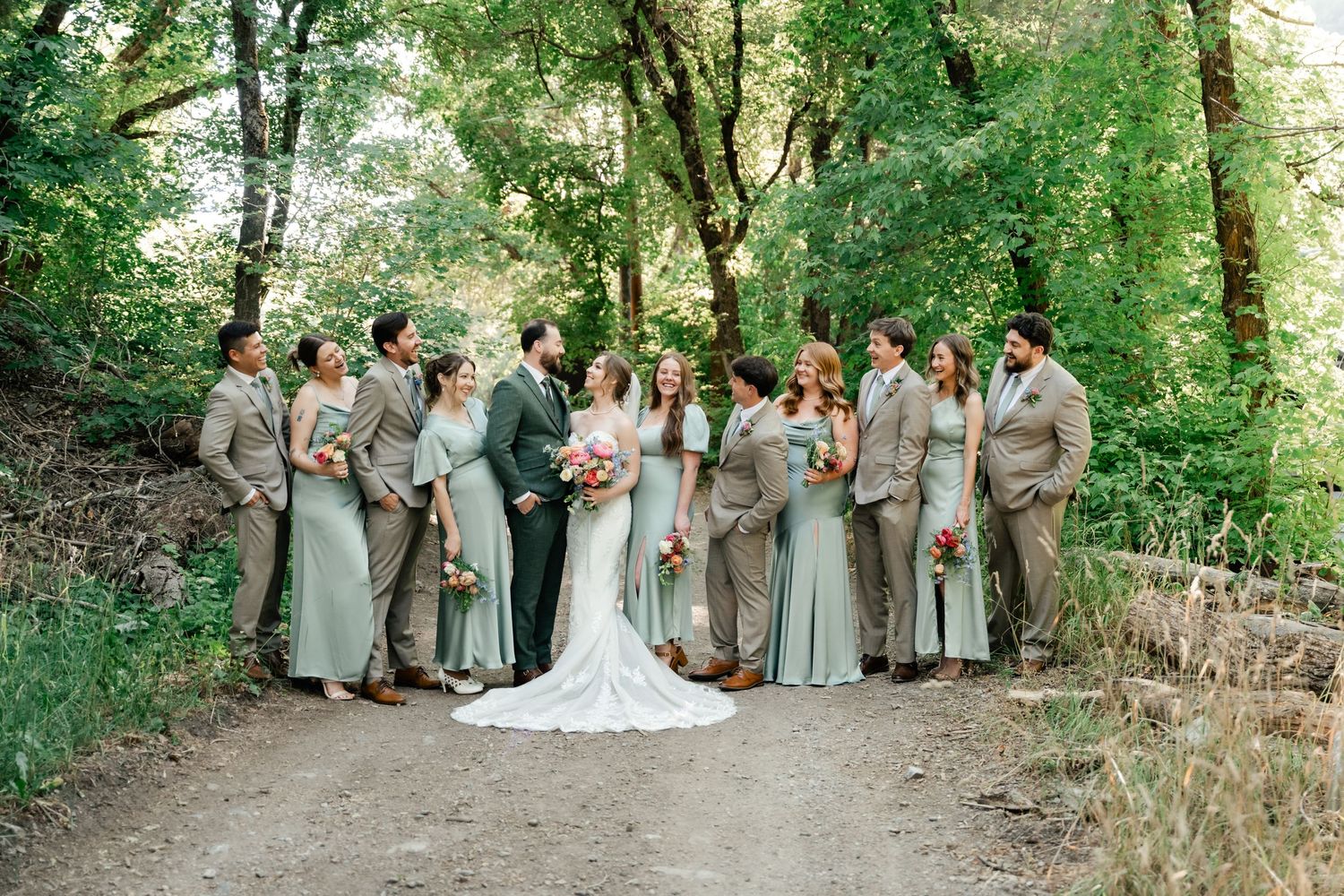 Wedding party in neutral attire poses together on a scenic wooded path surrounded by lush greenery. Millcreek Inn Wedding Photographer.