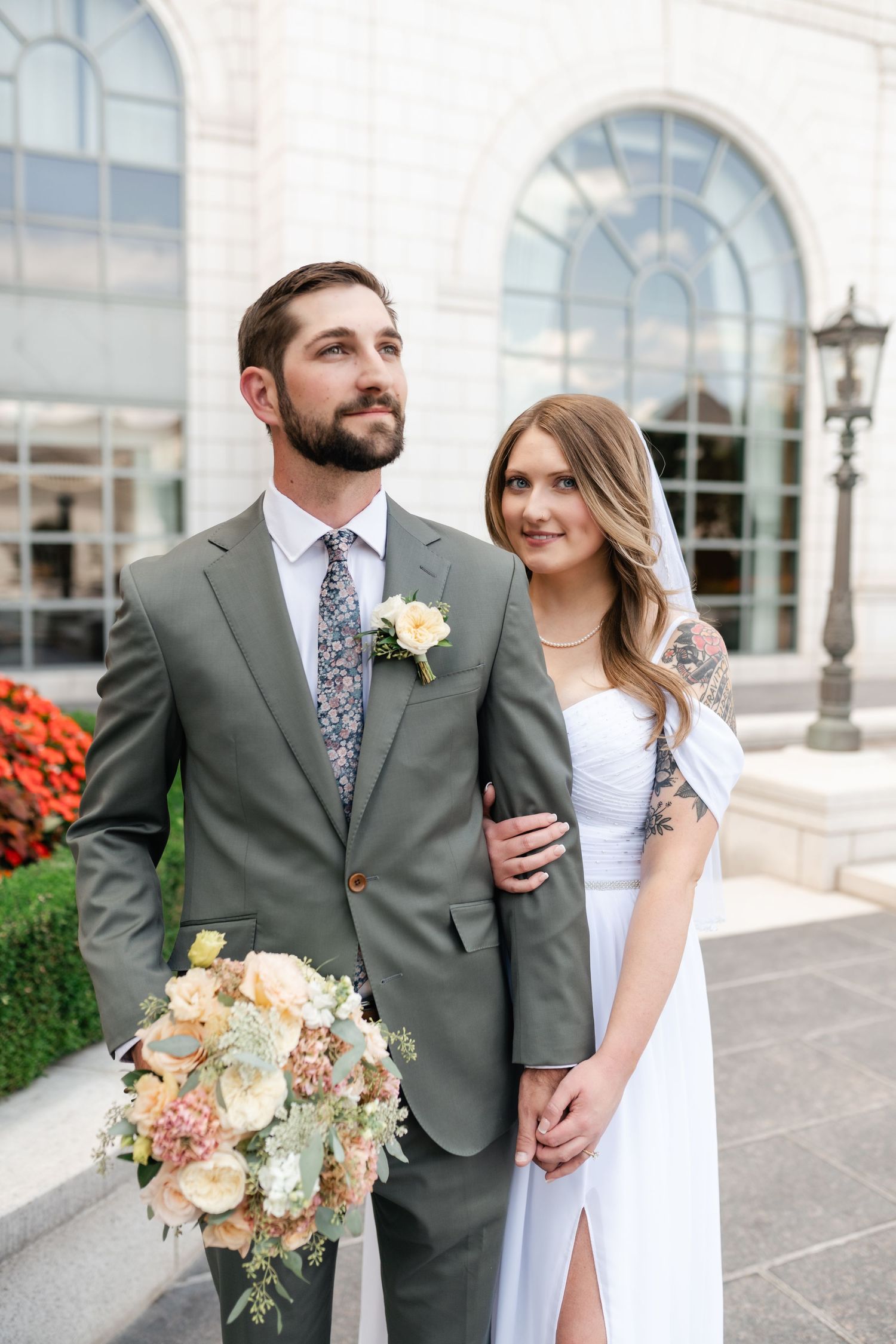 A couple in wedding attire pose together outside a building with arched windows and a peach-colored floral bouquet.