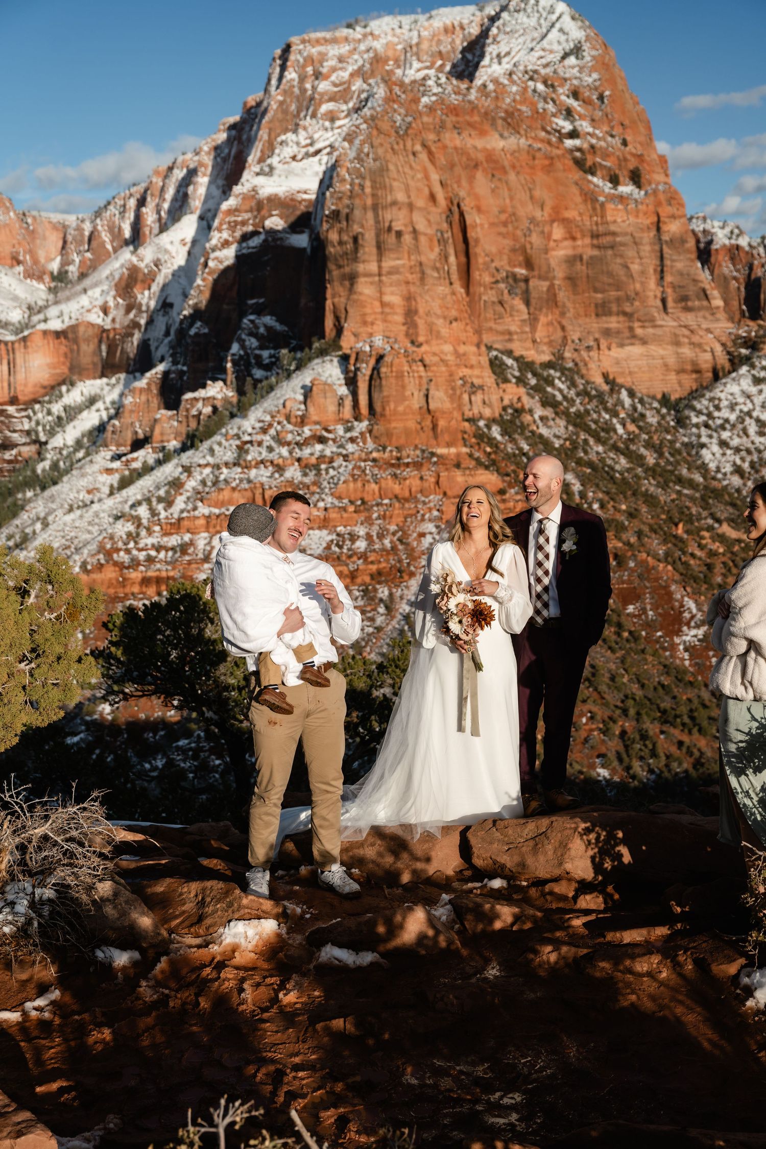A wedding ceremony takes place against dramatic red rock cliffs and snow-capped mountains.