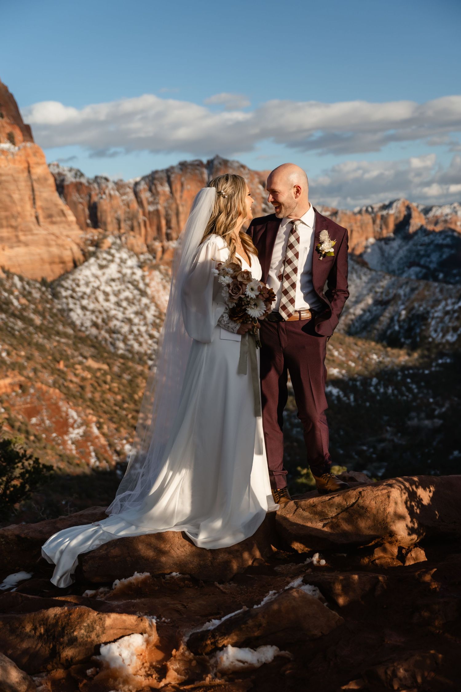 Wedding couple poses on red rock overlook with snow-dusted mountains in distance.