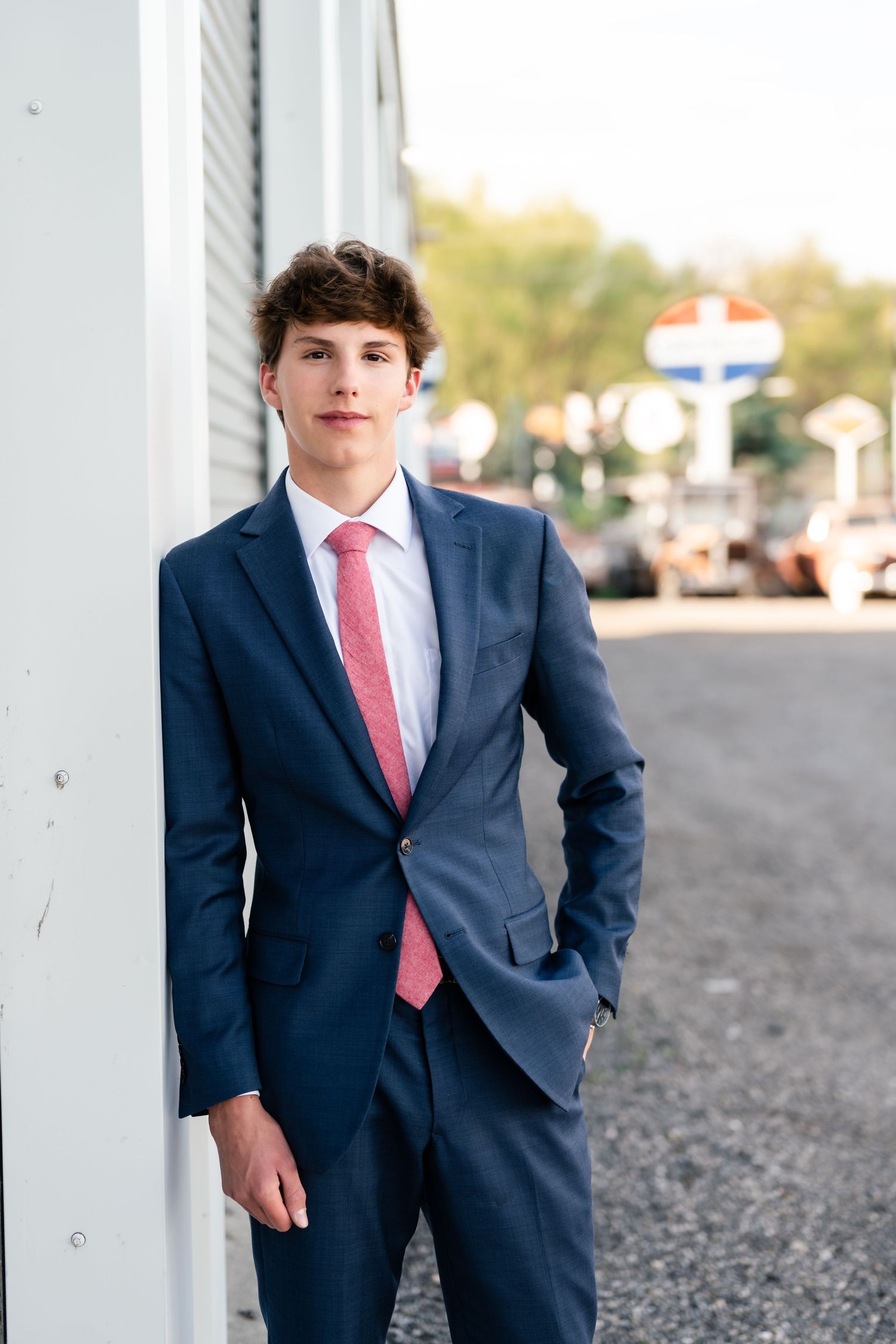 A person in a navy suit and coral tie poses against white columns on a gravel path.