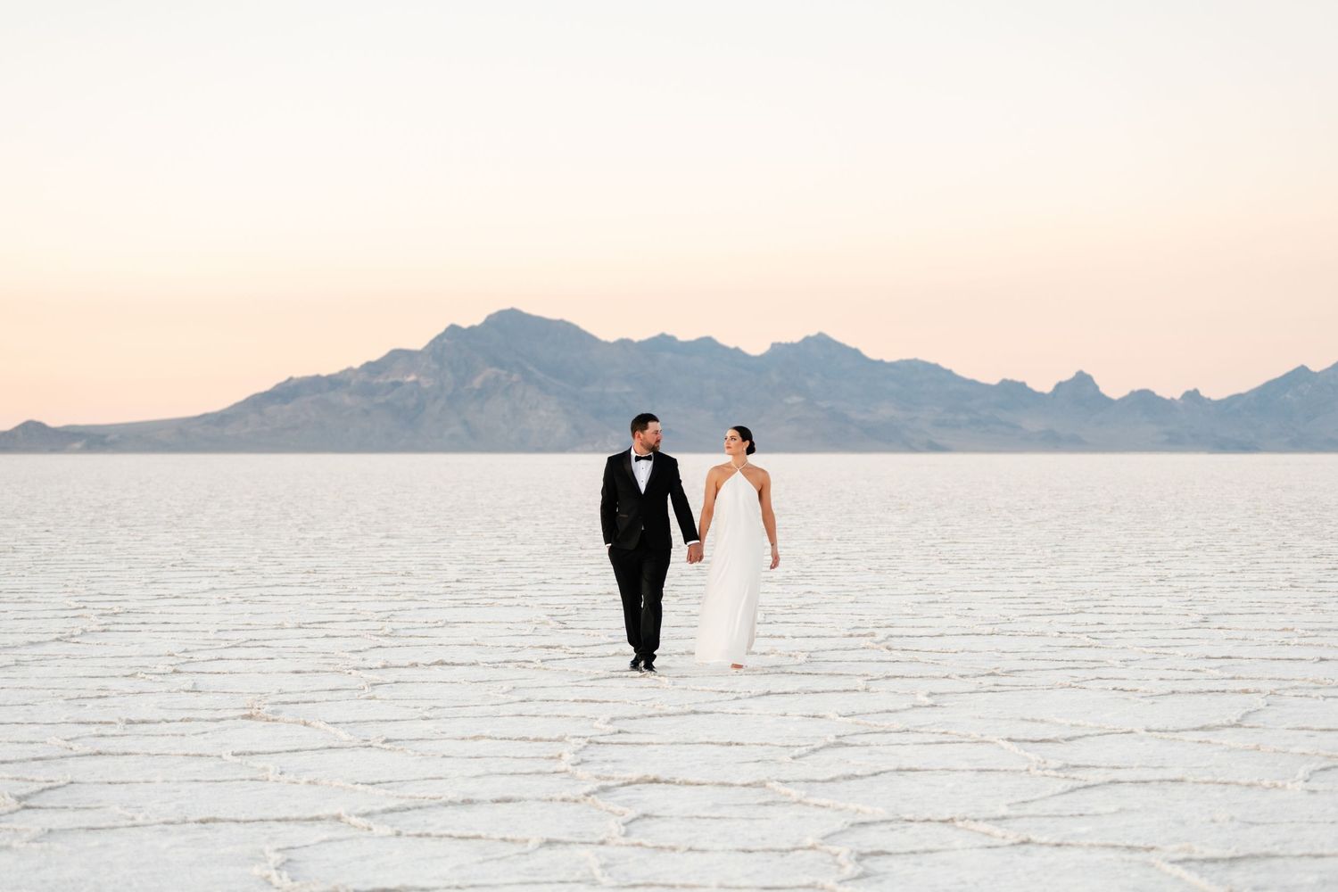 Utah Salt Flats Photographer. Elegant couple walks hand in hand across salt flats with mountain backdrop.