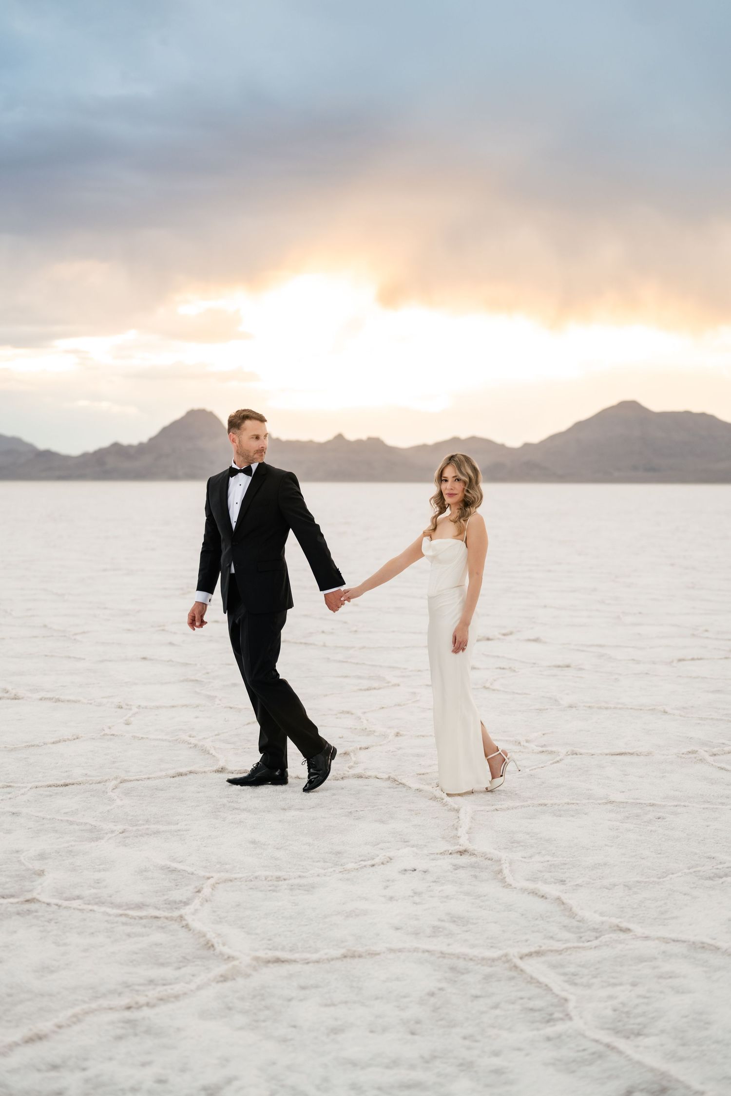 Salt Flats Elopement Photographer. Wedding couple walking together on cracked white salt flats with mountains and sunset sky in background.