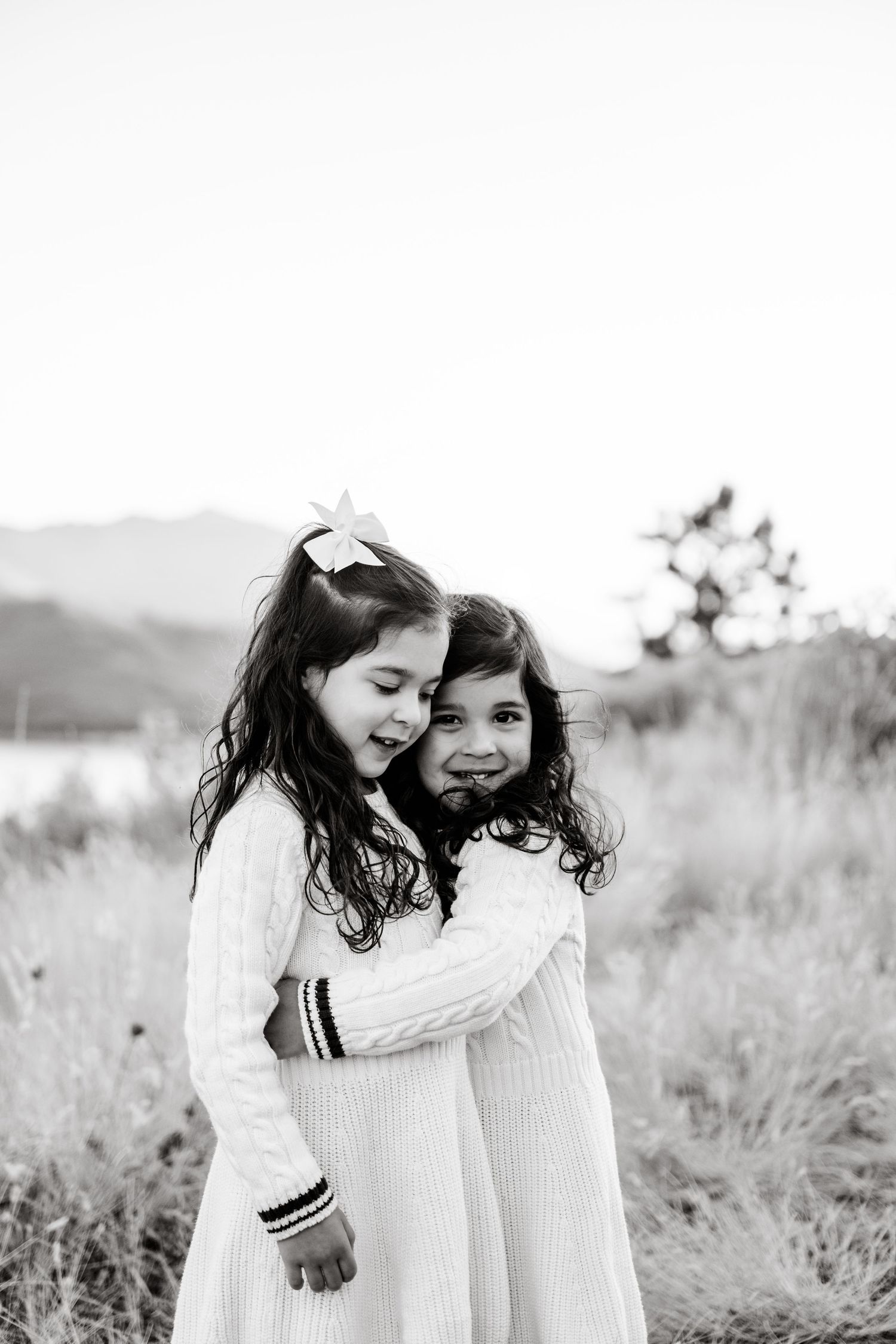 Black and white photo series showing two young girls in white dresses sharing sweet sisterly moments in a grassy field.