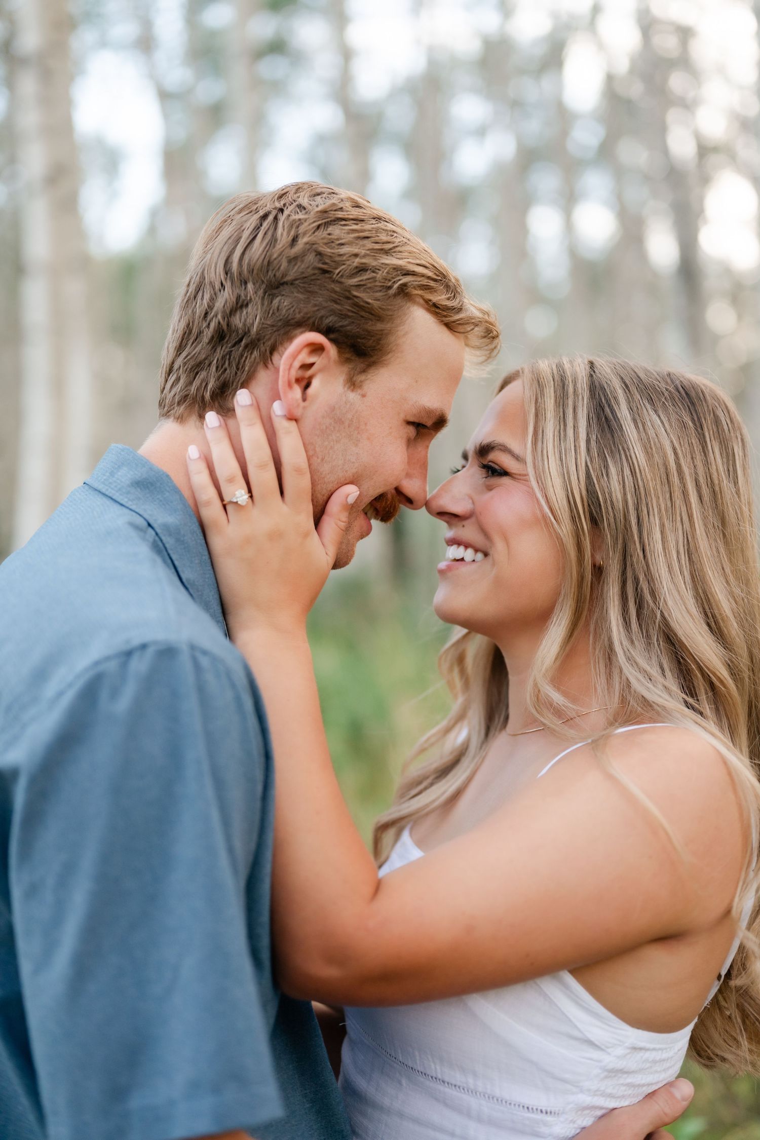 A couple shares a romantic nose-to-nose moment outdoors, smiling warmly, with the woman wearing an engagement ring.