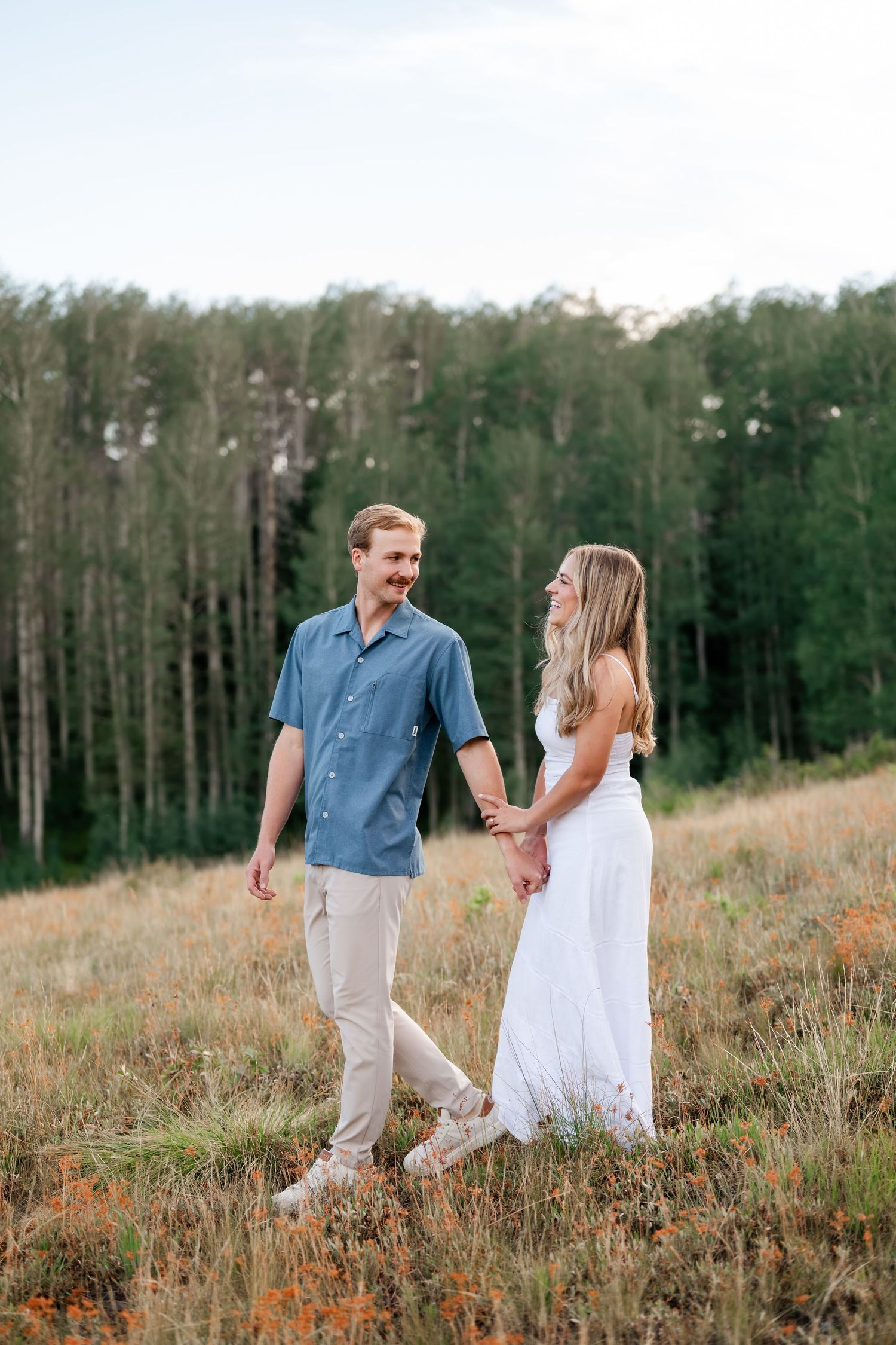 A couple holds hands walking through a wildflower meadow with tall green trees in the background at their engagements near Deer Valley.