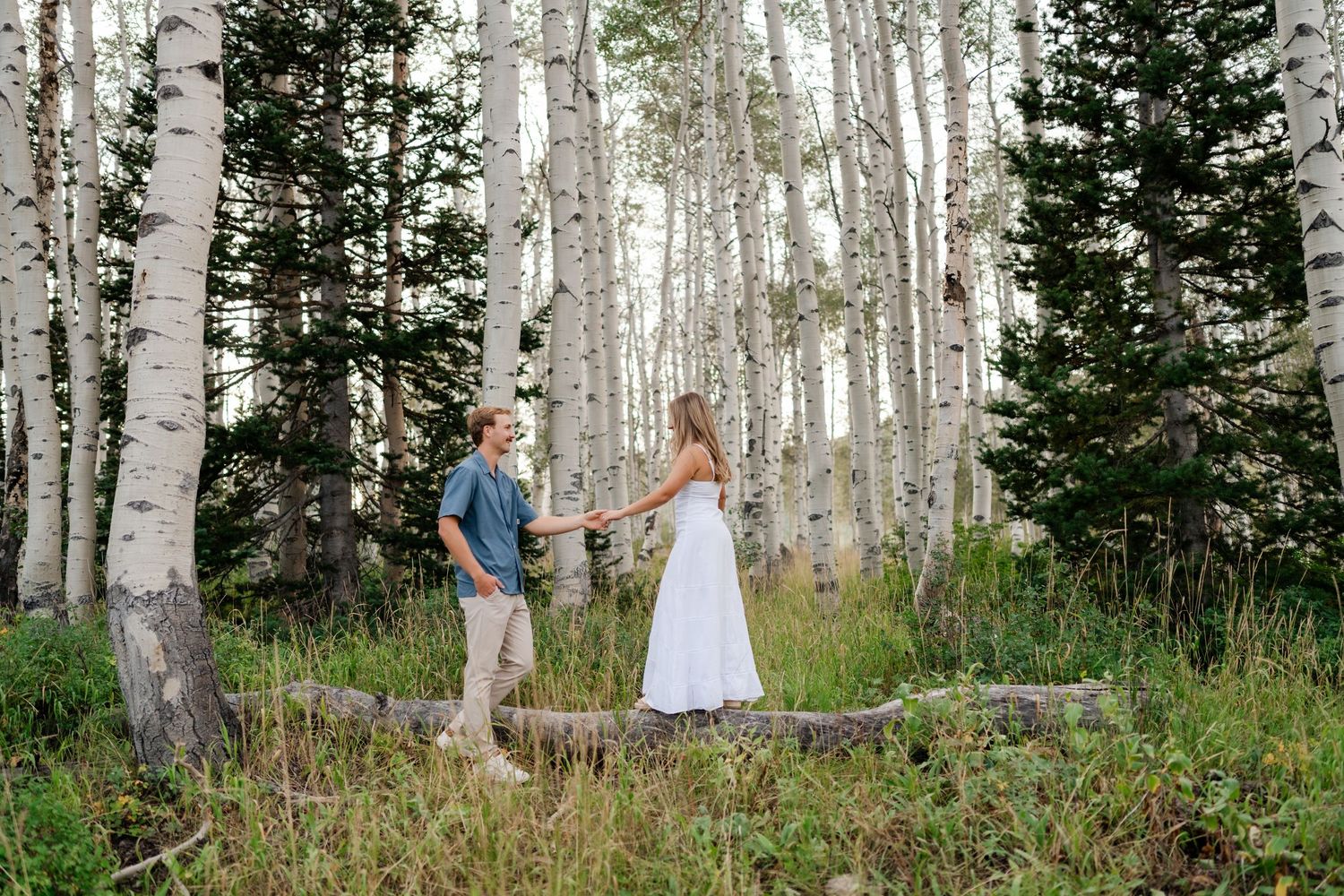Couple holding hands in a lush aspen forest, woman in white dress, man in blue shirt, surrounded by tall birch trees in Park City.