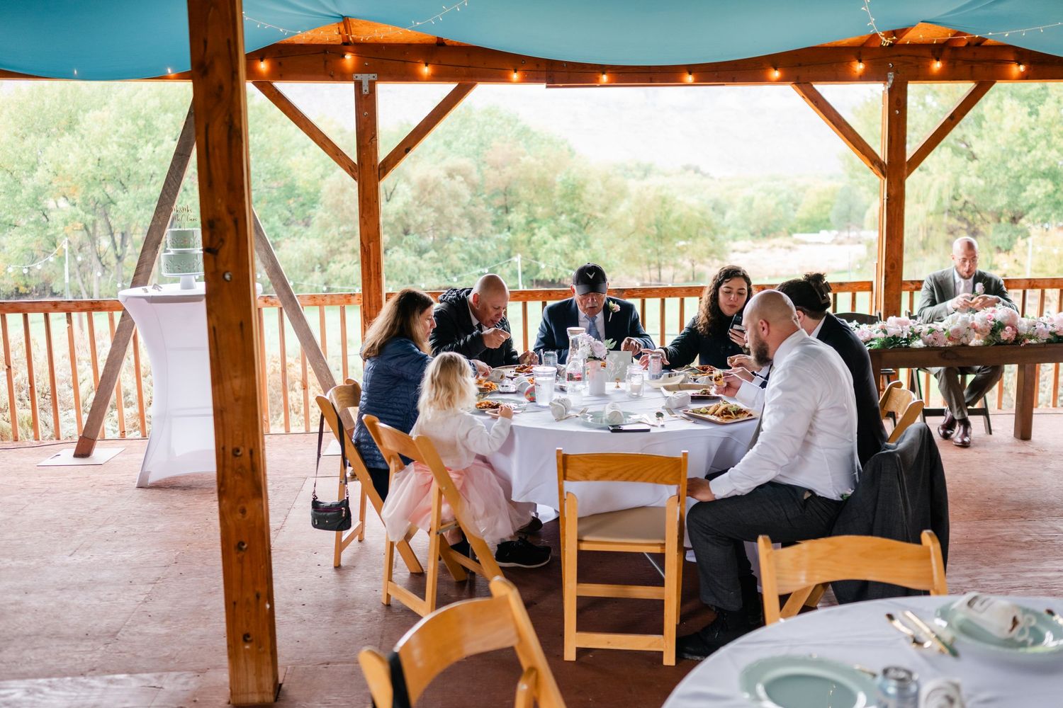Wedding guests seated at tables under a rustic wooden pavilion with string lights and scenic mountain views.
