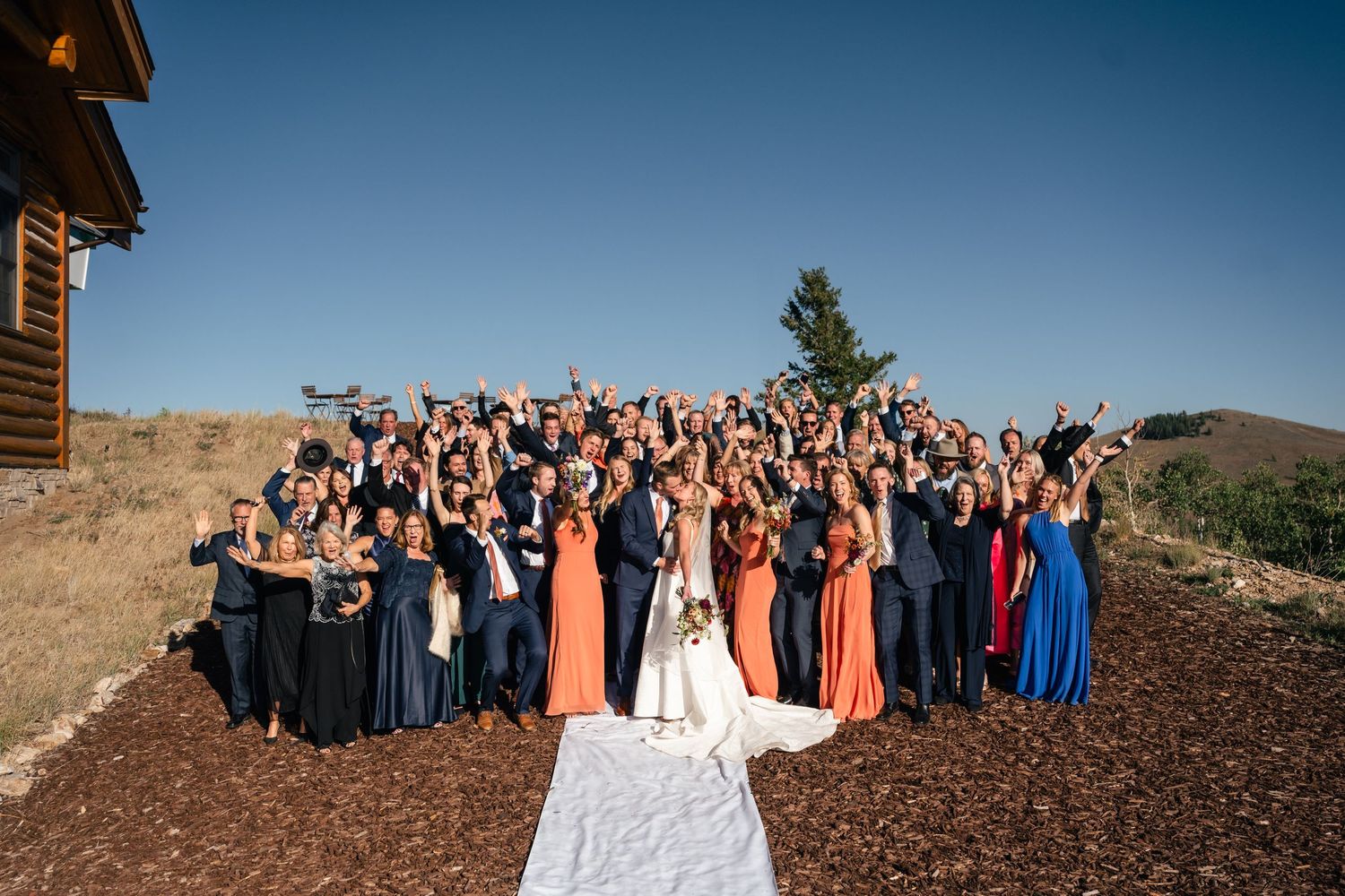 Large wedding party poses together on a scenic hilltop with mountains in background and long white dress train in foreground.