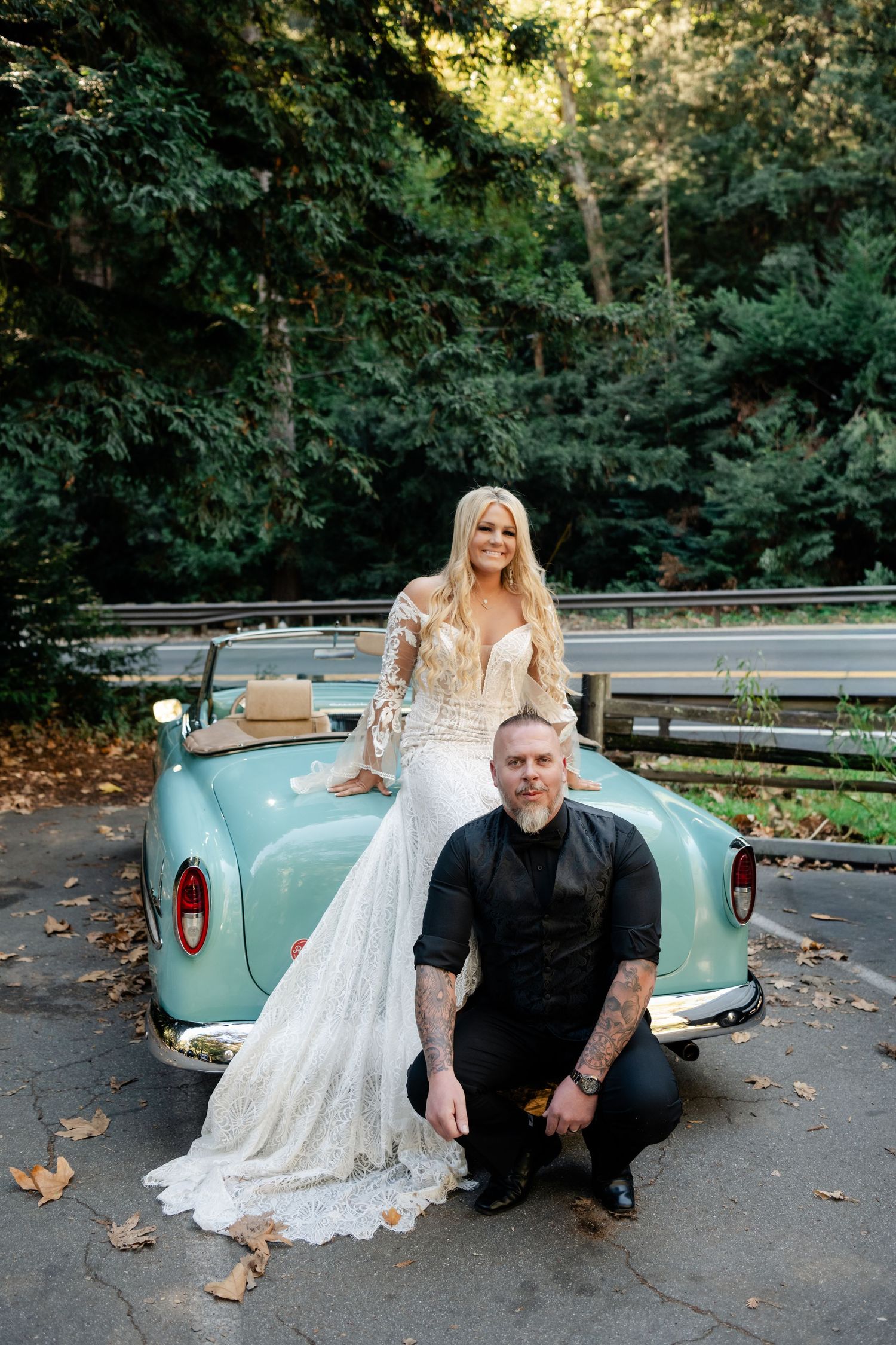 A wedding couple poses with a vintage mint green classic car surrounded by lush green trees.