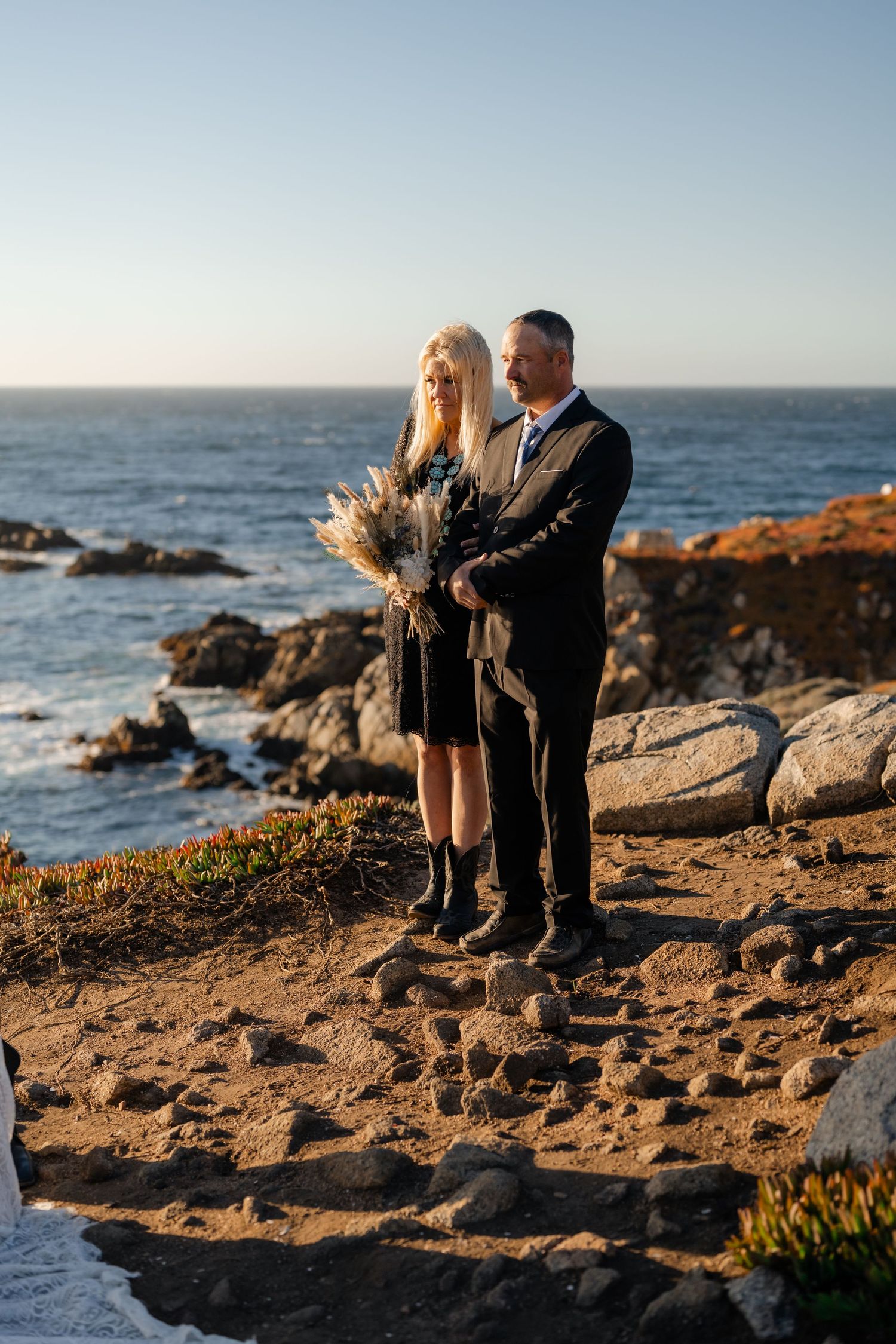 Newlyweds share a tender moment while seated on coastal rocks with ocean waves crashing behind them.