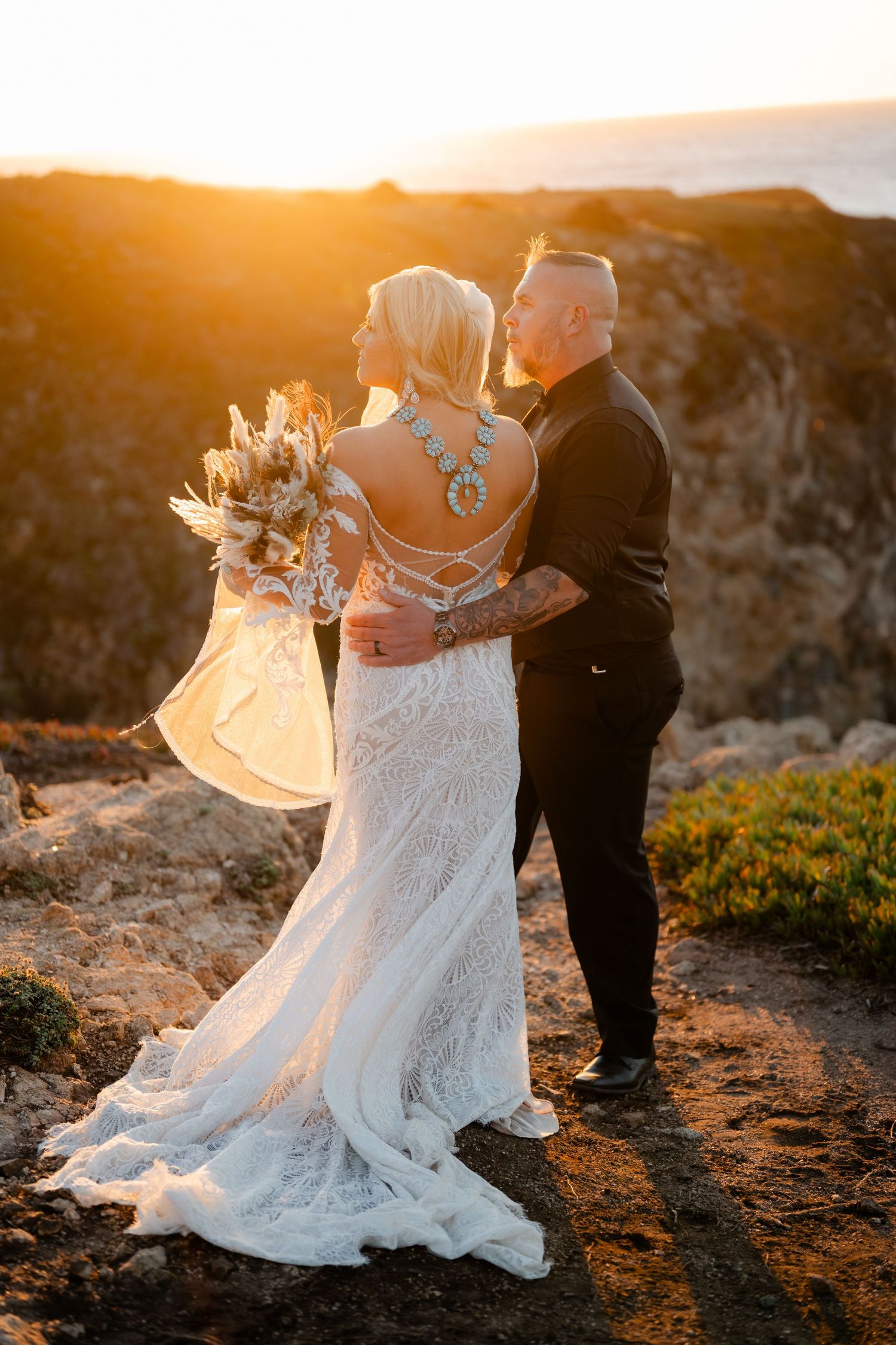 A romantic sunset wedding photoshoot shows a couple in formal attire dancing together on a cliff overlooking the ocean.