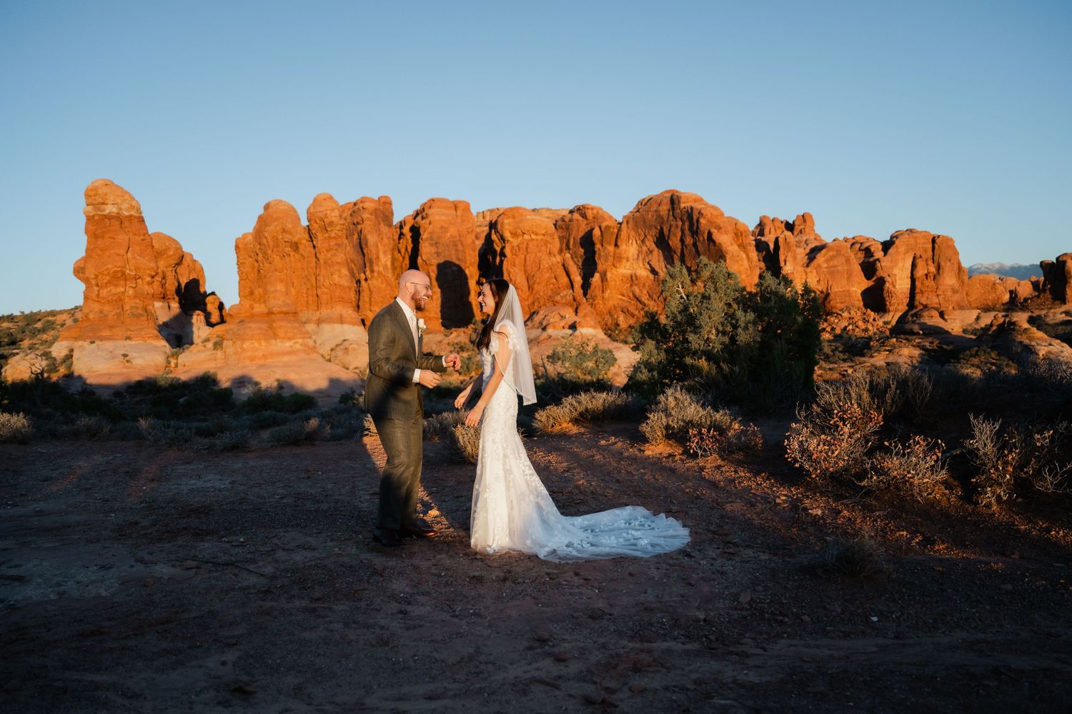 A couple in wedding attire poses among the stunning red rock formations at Arches National Park during sunset.