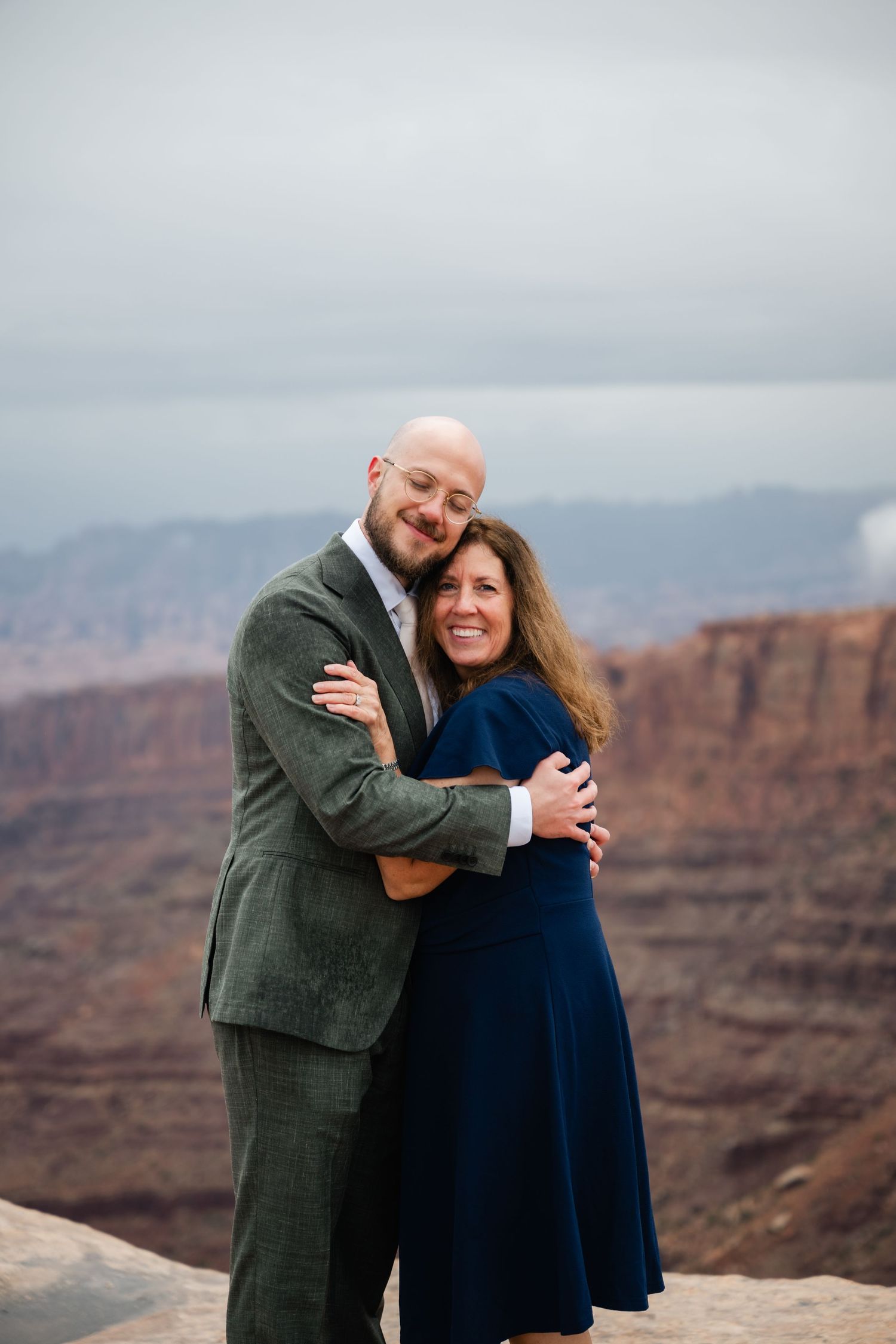 A couple embraces while taking photos at the edge of a dramatic canyon landscape on an overcast day.