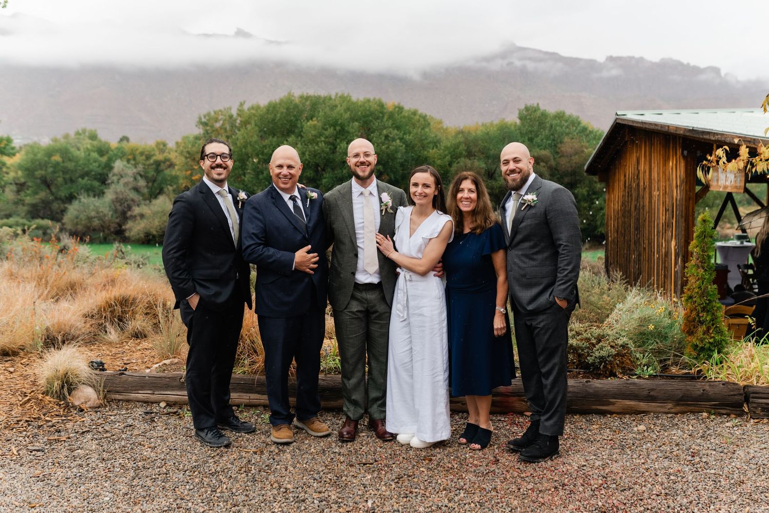 Wedding party members in formal attire gather for outdoor group photos in desert setting.