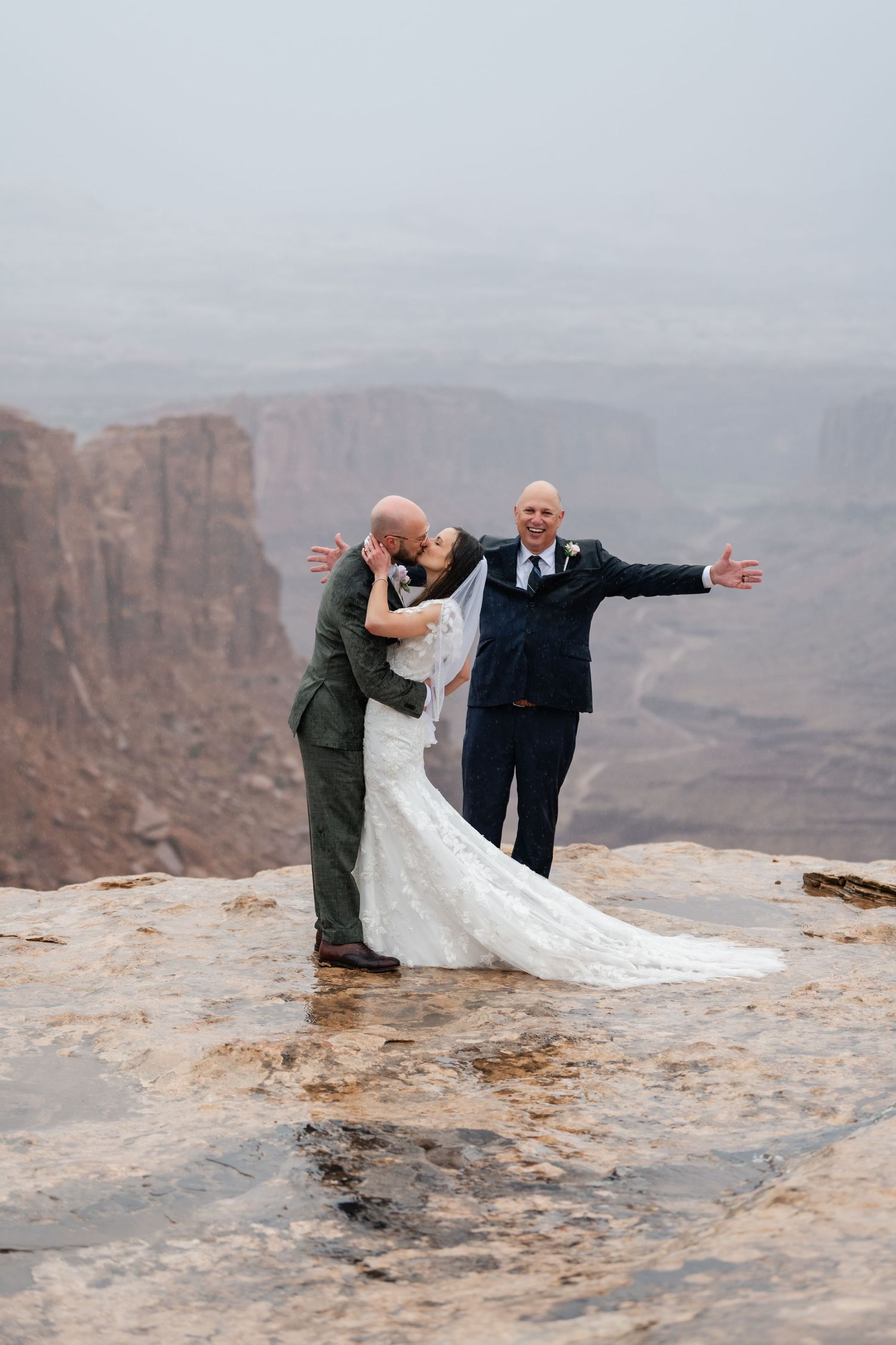Wedding couple and officiant stand at the edge of a cliff with flowing white dress billowing in the wind.