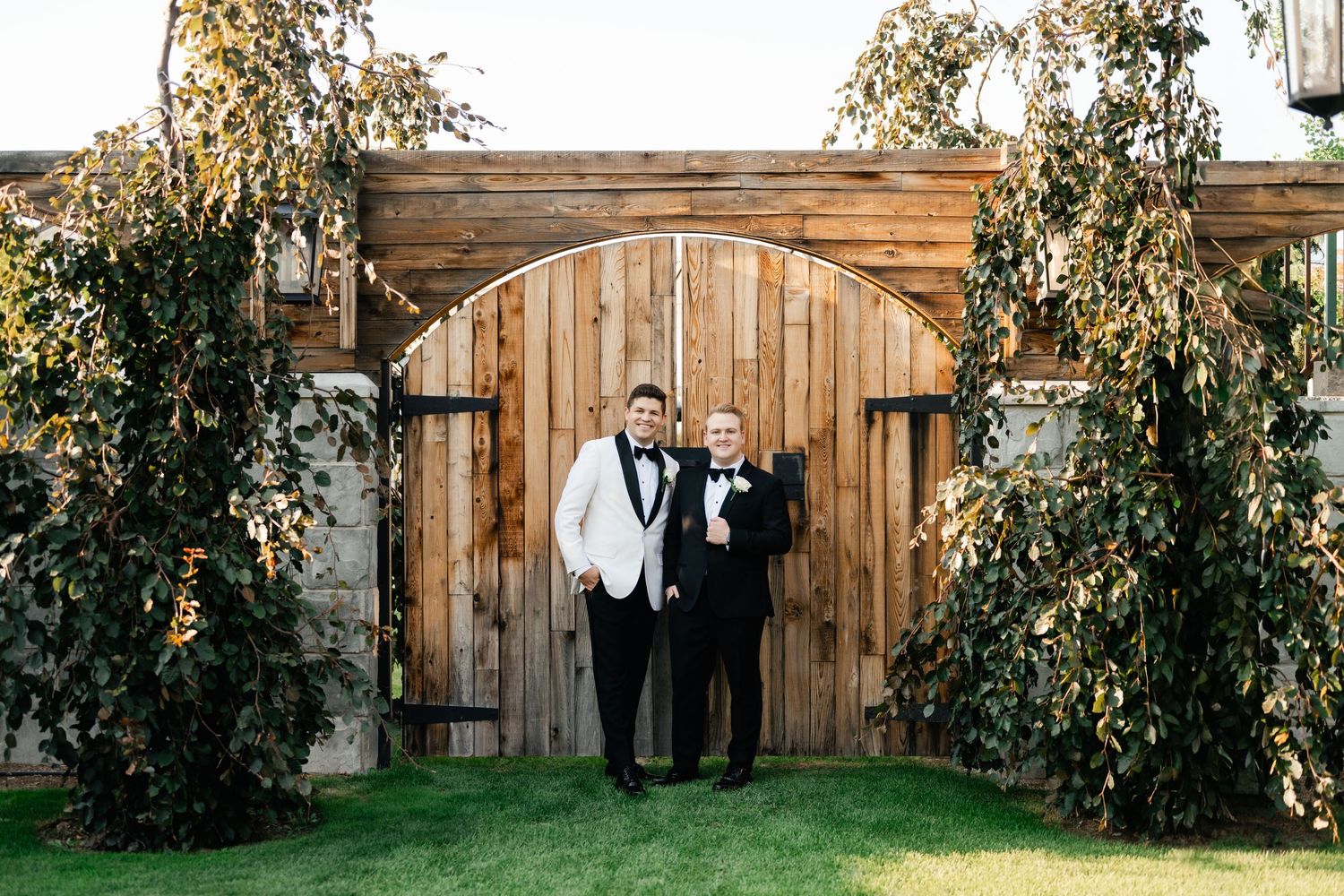 Two people in formal black and white attire stand in front of a rustic wooden gate with climbing vines at an Highland Gardens.