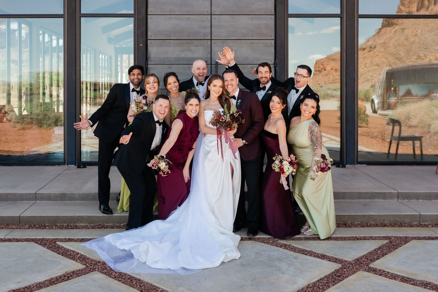 Wedding party poses joyfully outside a modern venue, bride in white gown, groom in burgundy, bridesmaids in green and wine dresses.