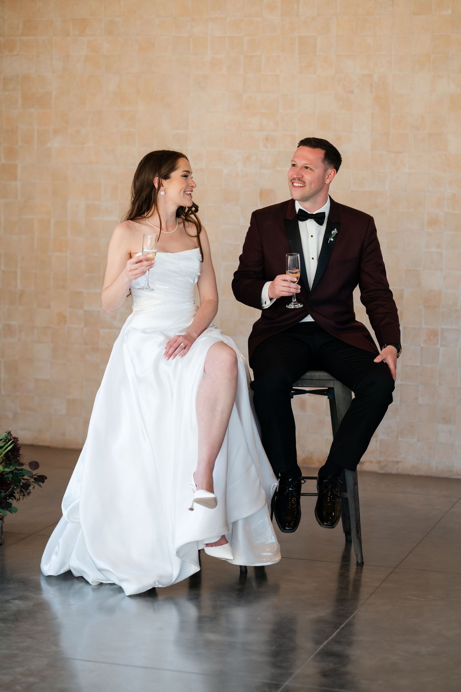 Bride and groom sitting on stools, smiling and holding champagne glasses, wearing white gown and burgundy tuxedo.