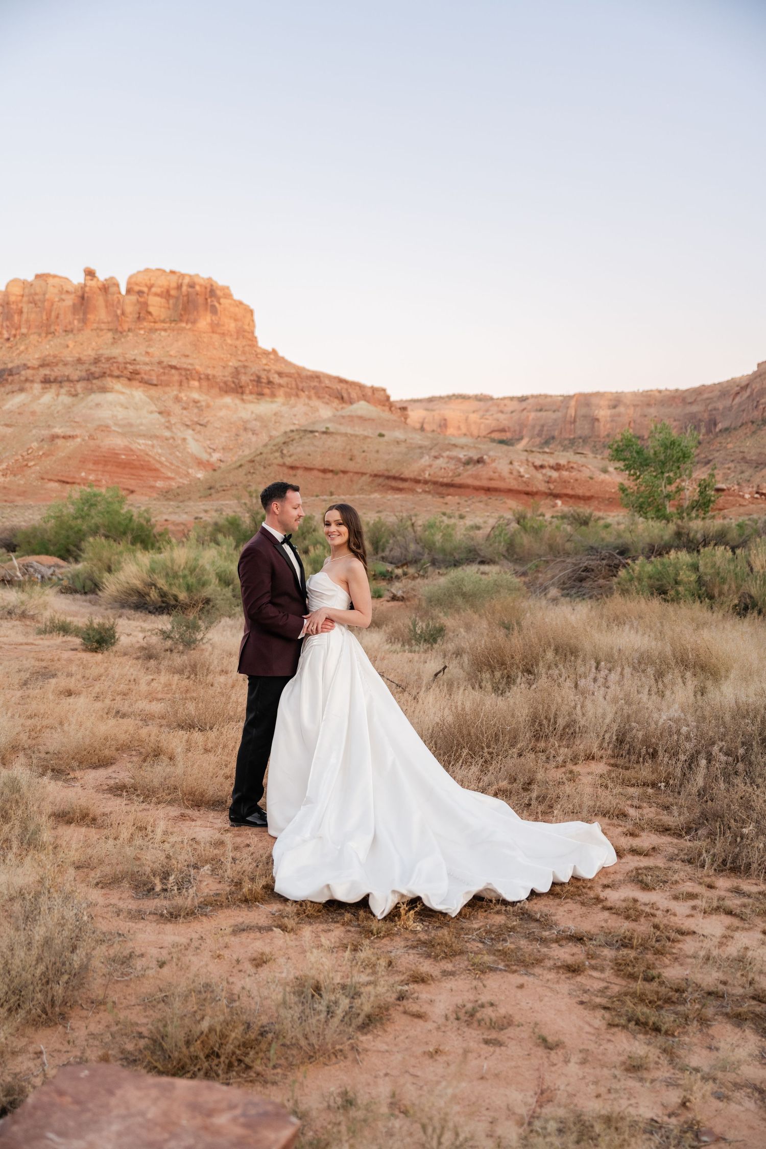 Bride and groom pose in white gown and burgundy tux against red rock canyon landscape at golden hour.