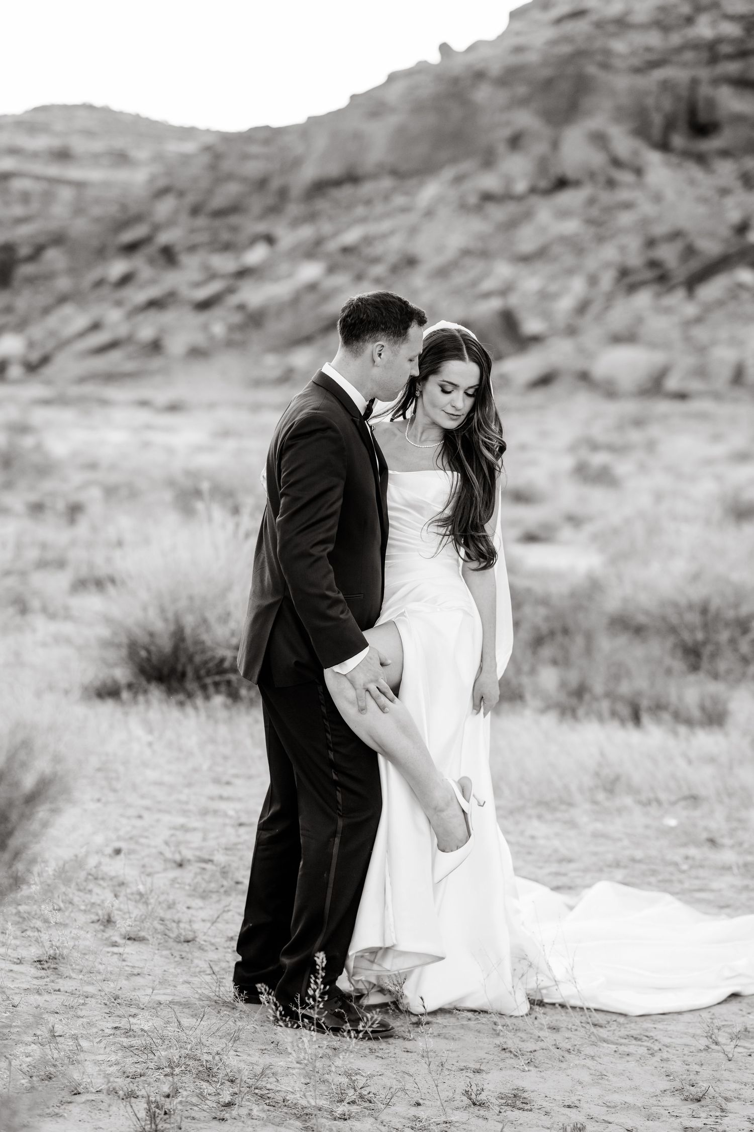 Black and white photo of bride and groom embracing in desert landscape with rocky mountains behind them.