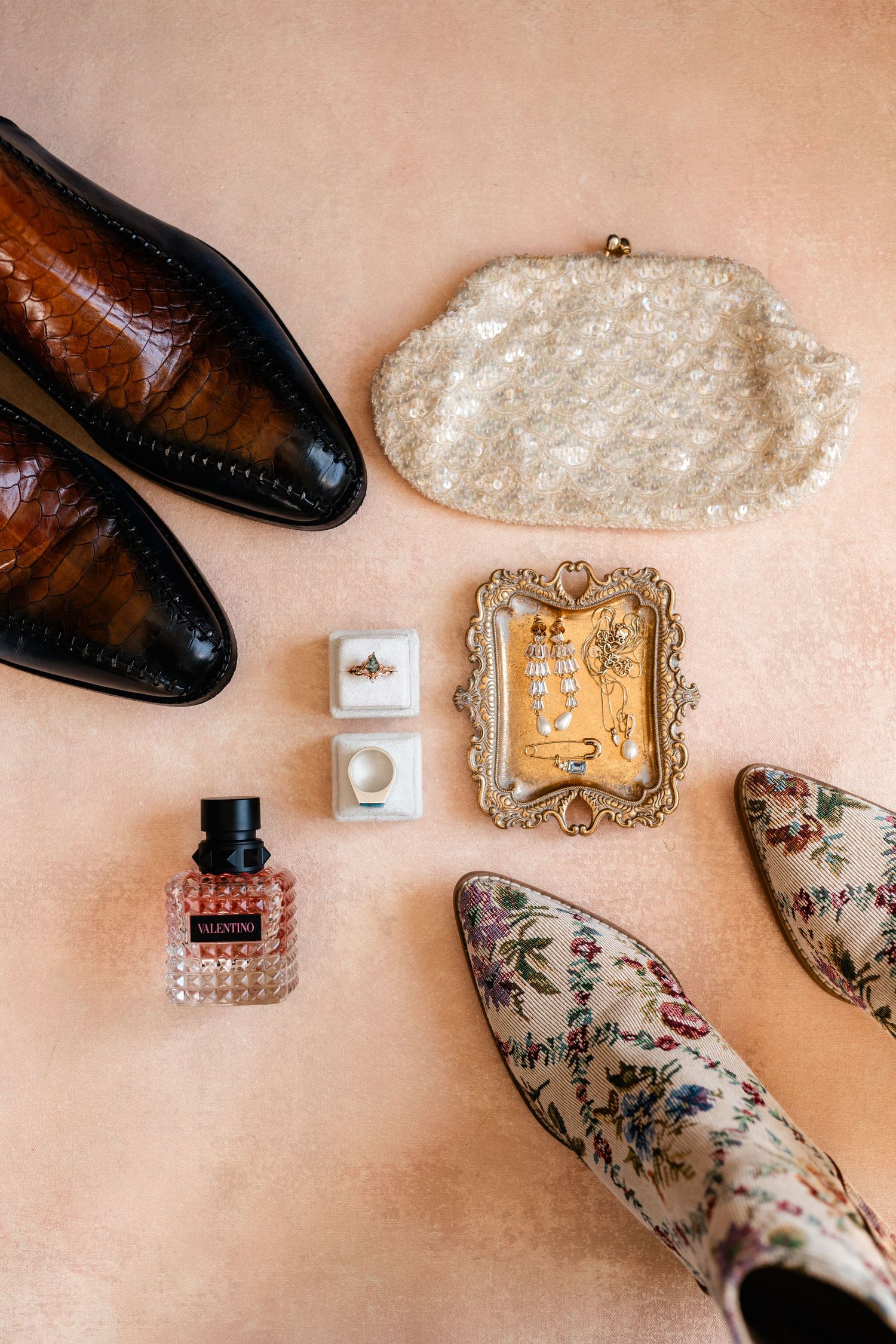 Wedding accessories including brown leather shoes, floral print shoes, perfume bottle and vintage frame on peach background.