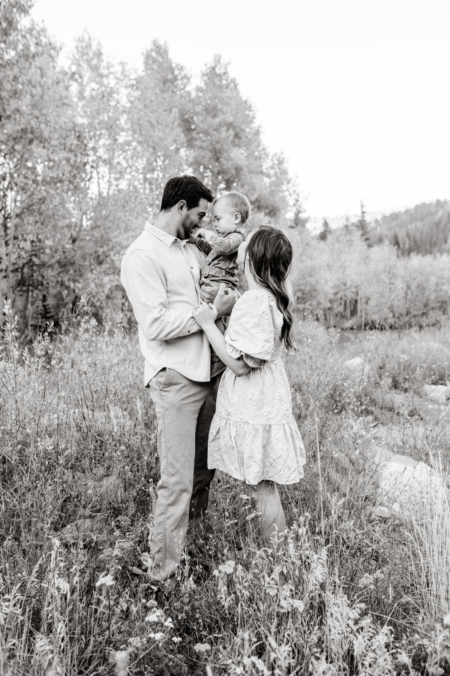 A couple shares an intimate moment with their child in a meadow with trees in the background.