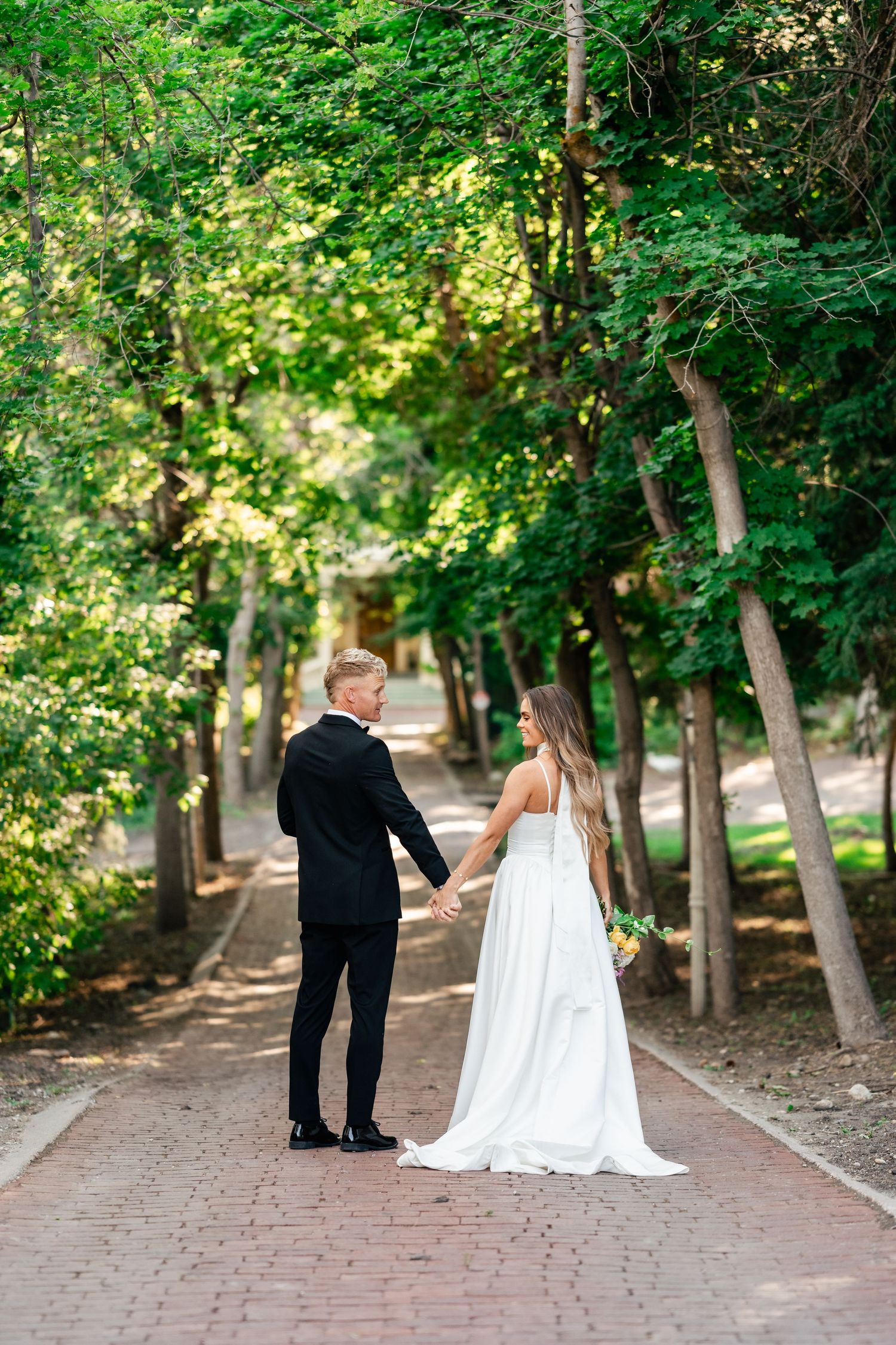 Couple walking down tree-lined brick path in wedding attire against forest backdrop.