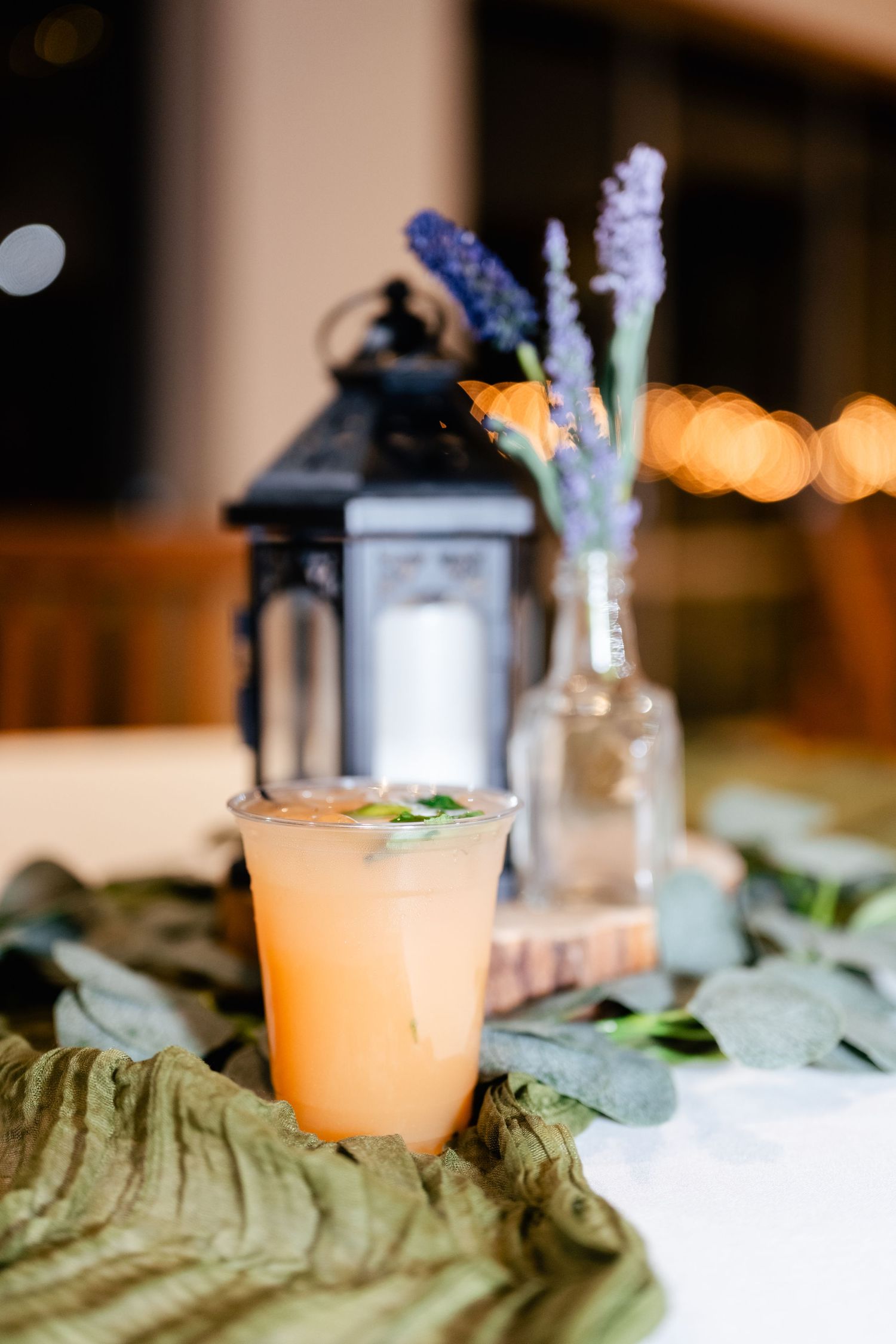 A peach-colored cocktail in a plastic cup sits beside a lantern and lavender stems on a draped fabric table setting.