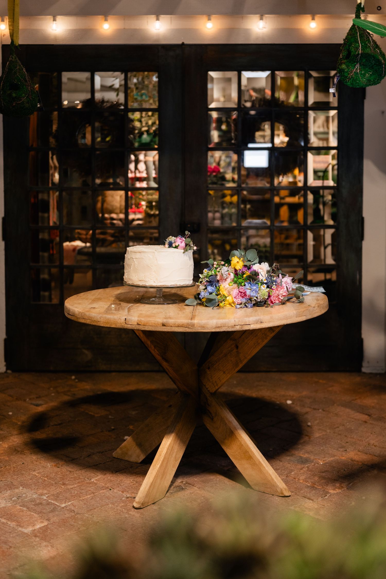 A white wedding cake and colorful floral arrangement sit on a rustic wooden pedestal table with dark shelving in background.